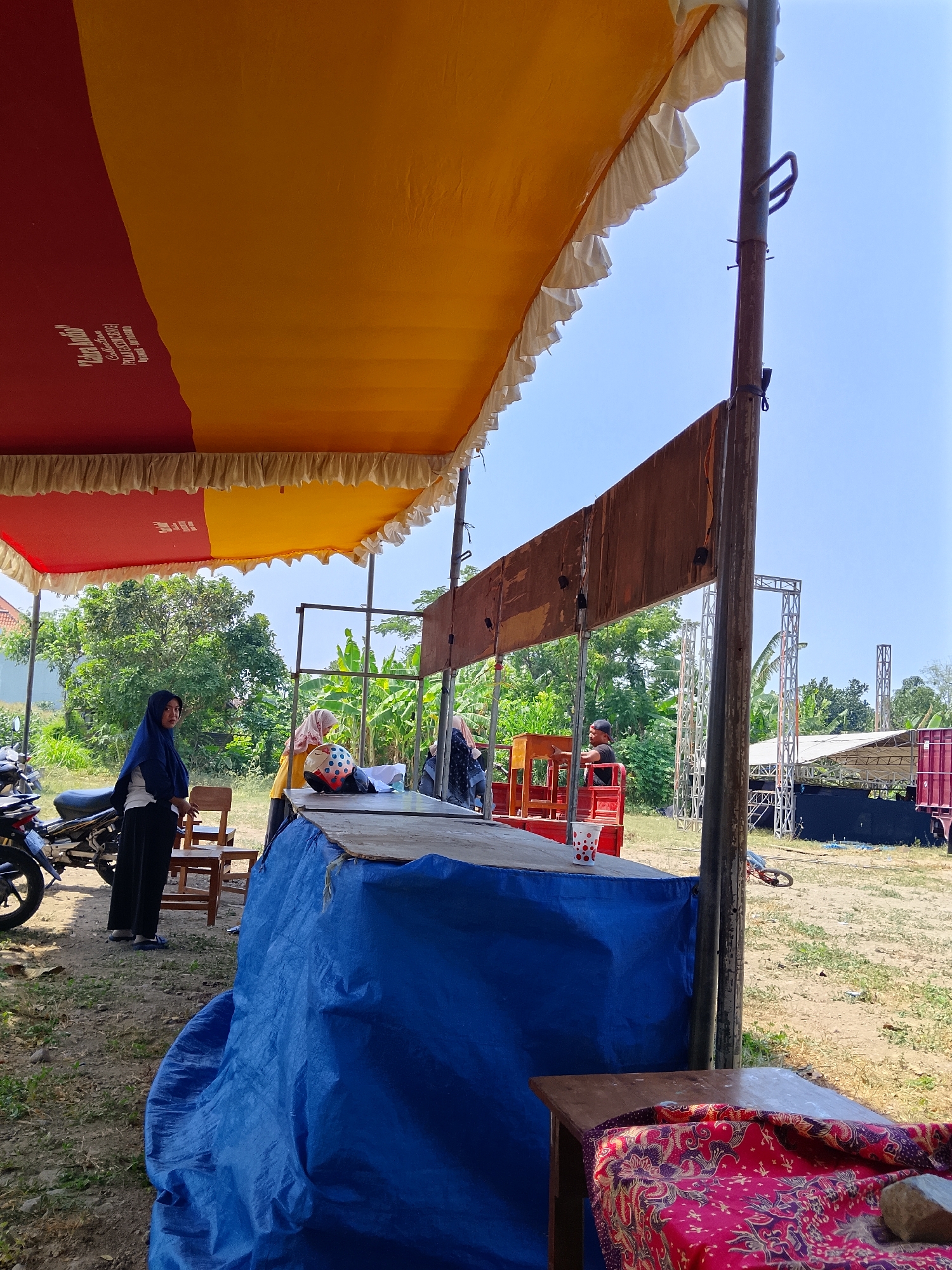 In the city of Pelem, Indonesia, an outdoor event appears to be set up. A large tent with red and yellow fabric provides shade, adorned with ruffled white trim. A person wearing a blue hijab and black pants stands next to a motorcycle, looking towards the left. Nearby, a wooden chair is visible. In the mid-ground, a raised platform is covered with a blue tarp on one side and a wooden surface on the other. Several individuals are positioned around this platform; one wearing a yellow top and a headscarf, another in a yellow top, and a third person in a black t-shirt and cap. A red structure with seating is also present, and a red and white polka-dotted cup sits on the platform. In the background, scaffolding and what appear to be stage lighting equipment are set up. Banana trees and other foliage are visible, suggesting a rural or semi-rural setting. The ground is covered with dry grass and dirt. A small table draped with a patterned red cloth is in the foreground on the right.