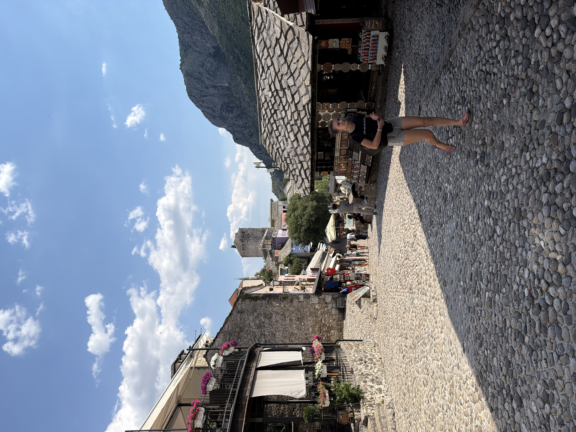 The image depicts a woman standing on a cobblestone street lined with shops selling souvenirs. She is wearing a black t-shirt with text that appears to say "EMPORIO ARMANI" and grey shorts, with pink flip-flops. Behind her, a mountainous landscape is visible, with a clear blue sky and scattered white clouds. The architecture suggests an old European town, with stone buildings and a paved walkway. Shops on the right display various trinkets and items, with some merchandise visible on shelves and tables. People are seen browsing and walking in the background, indicating a lively atmosphere. The lighting suggests it is daytime, with strong shadows cast on the ground, implying a sunny day. The overall scene evokes a sense of tourism and exploration in a historic setting.