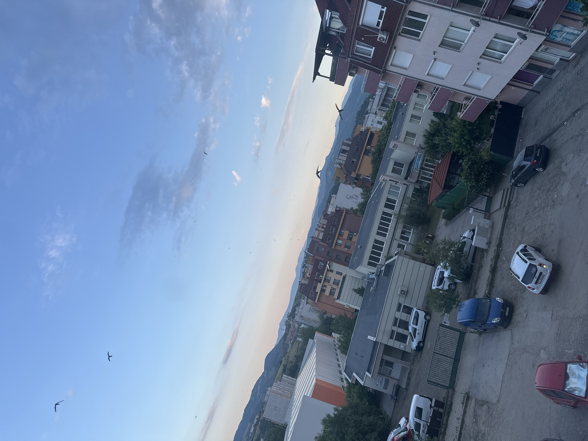 This image presents an elevated, oblique view of an urban environment in Blagoevgrad, Bulgaria, under a partially cloudy, blue sky. The perspective suggests it was captured from a multi-story building.
The upper portion of the image is dominated by a light blue sky with scattered, wispy white and grey clouds. Numerous small birds, consistent with swallows or swifts, are visible in various stages of flight across the sky, particularly in the upper left and right quadrants.
In the mid-ground, a range of green, undulating mountains forms the distant horizon, extending horizontally across the frame. Below the mountains, the cityscape unfolds. Prominent structures include:
- On the left, a large, rectangular building with white and orange paneling on its exterior and a flat roof. Adjacent to it, another white building features a large mural depicting a human face.
- Towards the center and right, multiple multi-story residential buildings are visible, characterized by varied facades (white, light brown, red), balconies with railings, and red-tiled roofs.
- Several smaller, utilitarian buildings with flat dark roofs and light-colored facades are interspersed throughout the urban fabric. One building on the mid-left features a sign with Cyrillic text, clearly identifying it as a "Детска градина" (Kindergarten). Another sign appears to be for a school ("ОУ" - "Основно Училище" - Primary School).
The foreground shows a paved area, likely a street or parking lot, exhibiting cracks and uneven textures. Numerous vehicles are parked or stationary:
- In the lower left, a red two-door car is partially visible.
- In the lower-center, a blue van (Fiat Doblo type) is parked, displaying a Bulgarian license plate `EA 4821 AX`, where "EA" is the code for Blagoevgrad.
- To its right, a white Dacia Duster SUV is parked, bearing a Bulgarian license plate `TT 2441 C` (Targovishte).
- Further right, a dark grey hatchback is parked, showing a Bulgarian license plate `C 4299 AH` (Sofia).
- Other vehicles, including a white van and a white SUV, are visible parked near a building entrance in the mid-left.
A dark green metal fence with a gate delineates a property in the mid-left, leading to a building with a light-colored facade and a dark, gently sloped roof. Interspersed among the buildings are various green trees and bushes. The overall lighting is soft, indicative of either early morning or late afternoon. No individuals are explicitly visible.