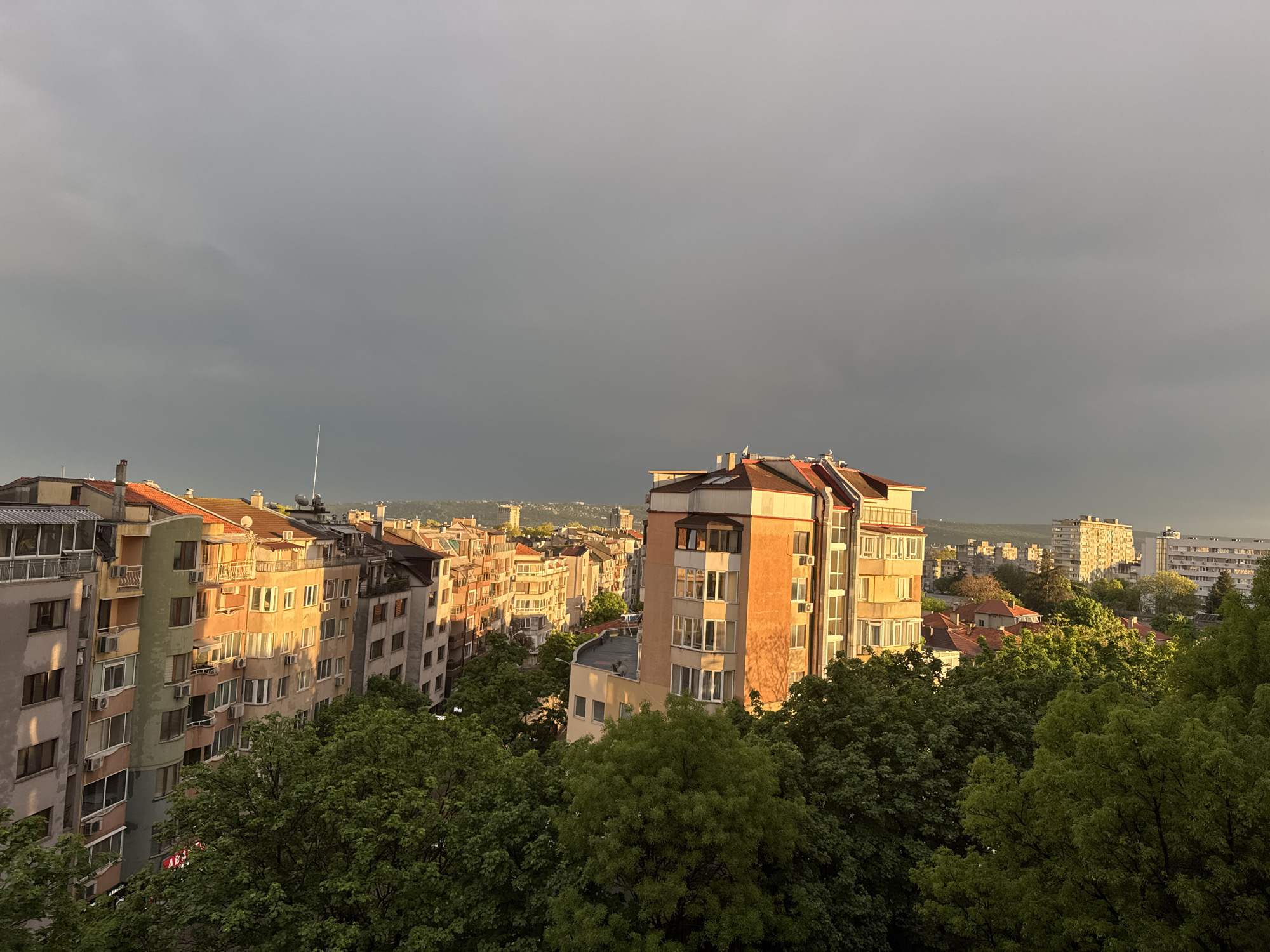The image shows an urban landscape in an unknown city, taken from an elevated perspective. The sky is predominantly grey and overcast. Numerous buildings, varying in height and design, occupy the scene, with some trees visible in the foreground. The sunlight appears to be breaking through in patches, casting a warm glow on certain buildings. A lightning rod extends from the roof of one of the buildings.