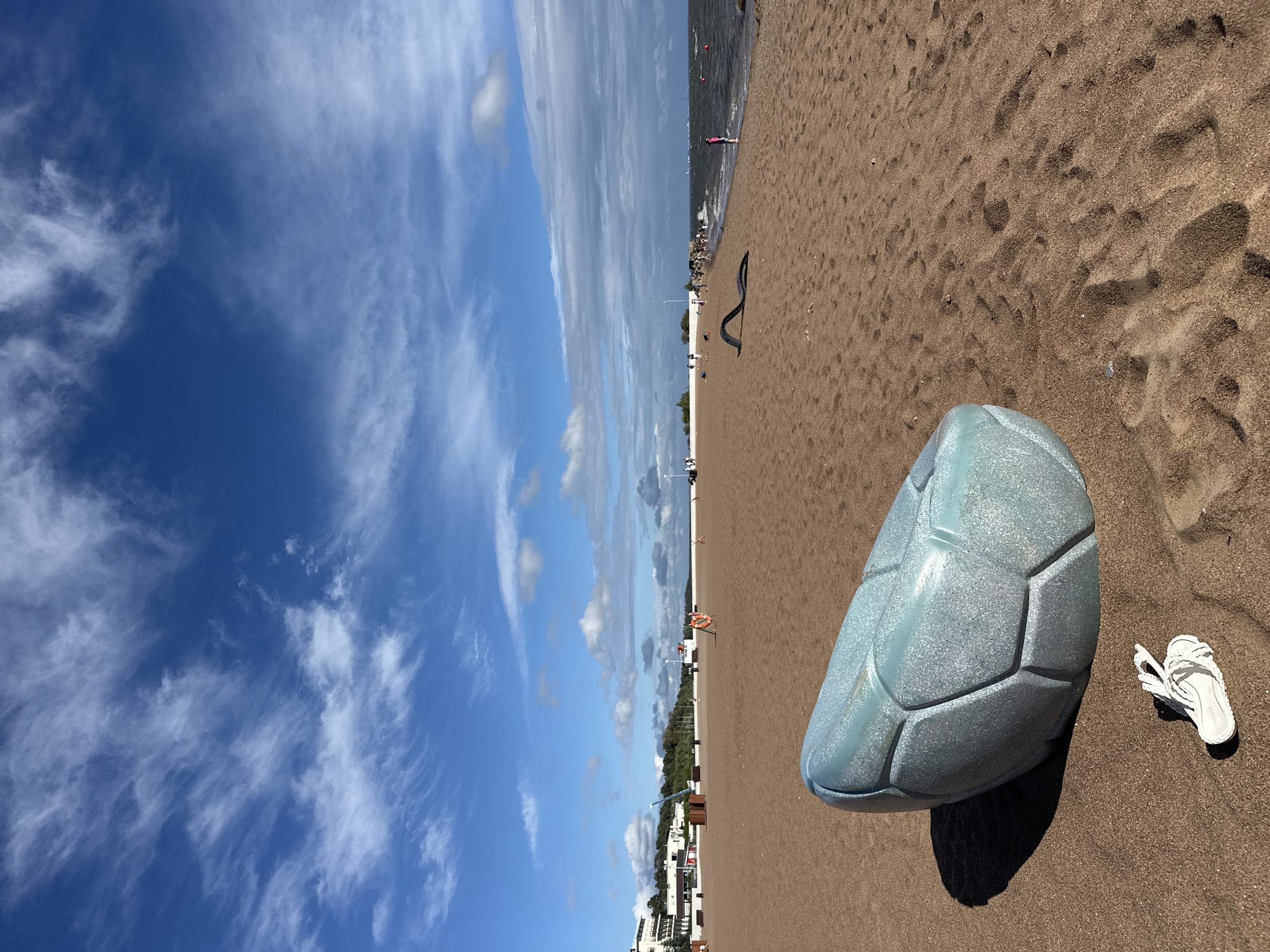 The image depicts a sandy beach scene in Zelenogorsk, Russia, under a partly cloudy sky. In the foreground, a large, light blue, geodesic-shaped object resembling a soccer ball is positioned on the sand. Next to it, a single white sandal with braided straps lies on the sand. The beach stretches towards the background, where people are visible walking and standing near the water. The sea is calm with moderate waves. Along the shoreline, there are various amenities, including what appear to be benches, a life preserver, and possibly some recreational structures. Buildings are visible in the distance behind the beach. The overall environment is that of a public beach on a bright, possibly overcast day.