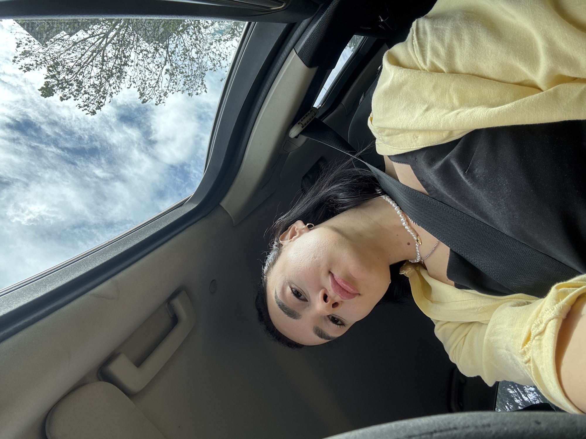 A woman with dark hair, wearing a black top, a yellow cardigan, and a pearl necklace is in a car. She's wearing a seatbelt and looking at the camera. The car has an open sunroof revealing a view of trees and a cloudy sky. The location is in Tbilisi, Georgia, as evidenced by the tree species commonly found in the region and the style of the car's interior.