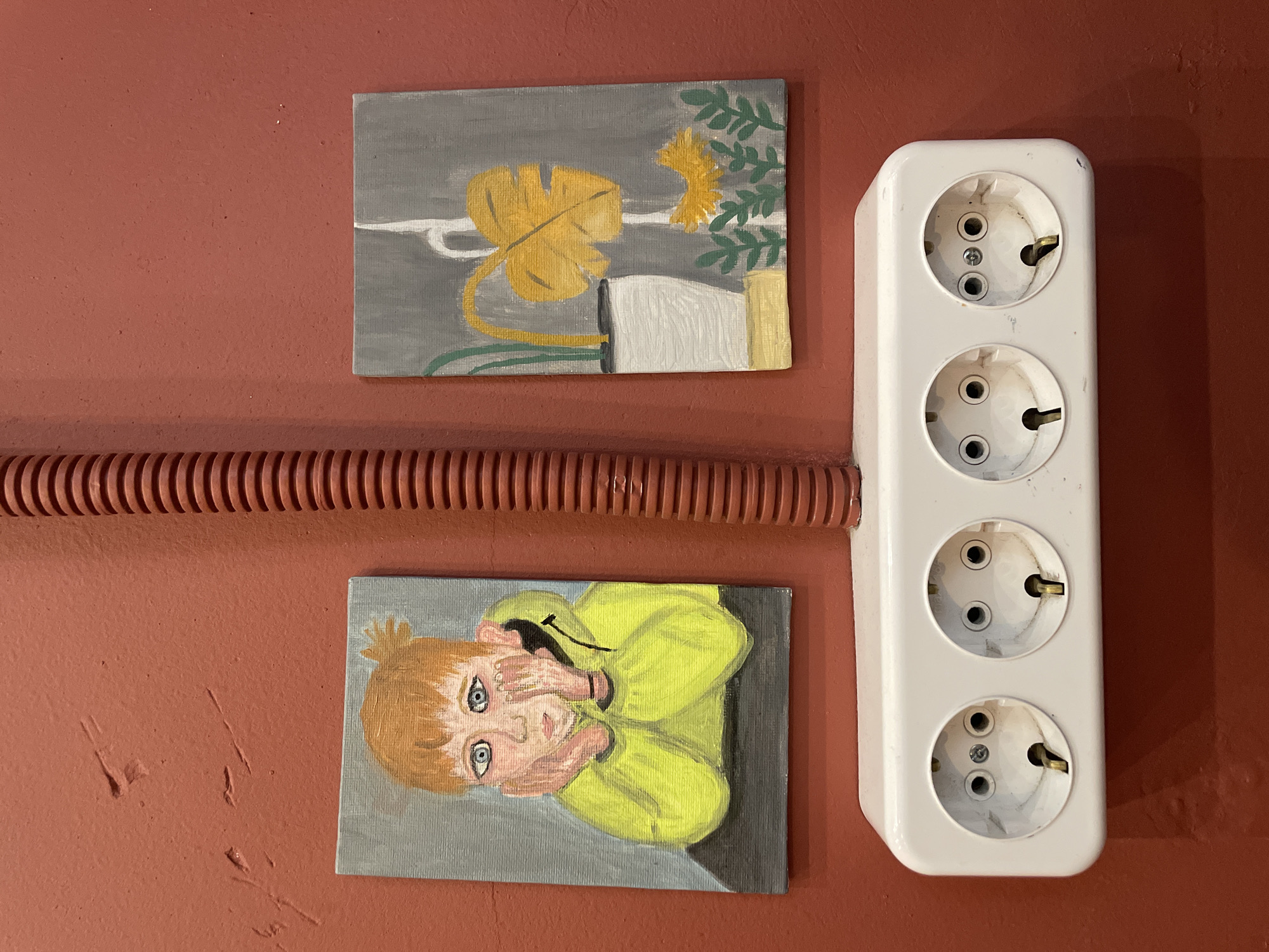 The image depicts a section of a wall with a reddish-brown hue. A corrugated brown pipe runs horizontally across the center of the frame. Above the pipe, a small painting shows a stylized depiction of a monstera leaf and a yellow flower in a pot on a gray background. Below the pipe is another painting featuring a person with fair hair wearing a yellow garment, their hands partially covering their face, set against a gray background. To the right of the paintings is a white electrical outlet with four sockets. The environment suggests an interior space, possibly a room in a building in Khimki, Russia, given the prompt. The scene includes paintings, an electrical outlet, and a corrugated pipe fixed on a painted wall.