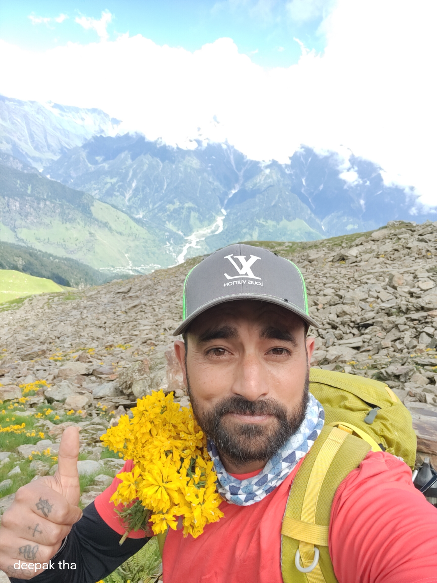 In a close-up, low-angle selfie taken from a mountainous region, a man wearing a red shirt and a grey baseball cap with a white "LV" logo is featured. He has a beard and is looking towards the camera. A garland of bright yellow flowers is draped around his neck. He is giving a thumbs-up gesture with his left hand, which displays several tattoos. Behind him, a green backpack is visible. The background shows a steep, rocky slope dotted with yellow flowers, leading to a vista of majestic, cloud-capped mountains. A faint inscription "deepak tha" is present in the bottom left corner of the image. The location is identified as the city of Bashisht, India.