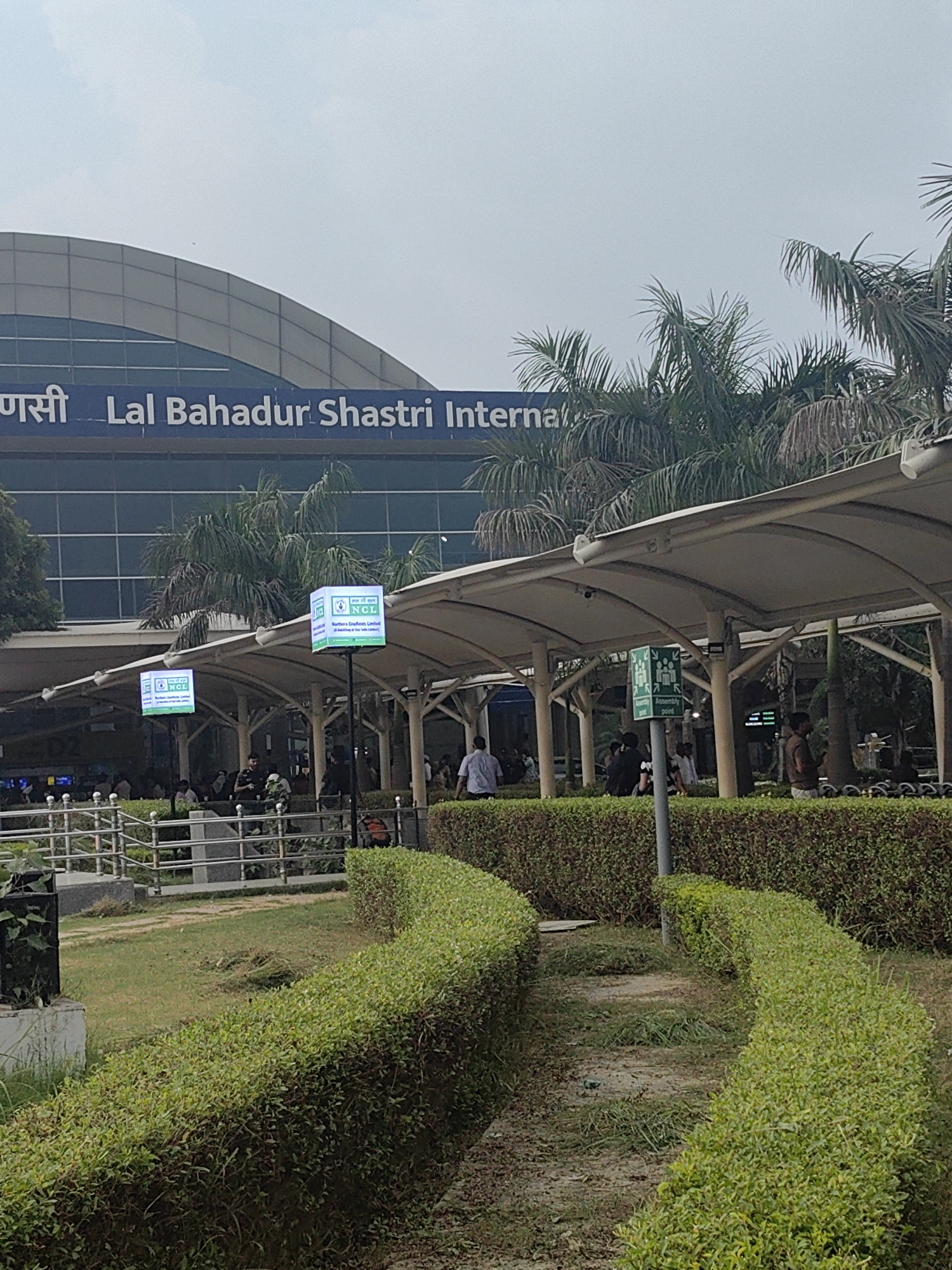 The image depicts the exterior of Lal Bahadur Shastri International Airport in Pura Raghunathpur, India. The airport building, with its blue facade and curved roofline, is visible in the background. In the foreground, a covered walkway with white canopy shelters leads towards the entrance. Several people are seen walking and standing under the walkway and in the surrounding area. There are well-maintained green hedges and a grassy area in front of the walkway. Two illuminated display boards are mounted on poles, showing company logos. A sign indicating an "Assembly Point" with pictograms of people is also visible. Palm trees are interspersed throughout the scene, indicating a tropical or subtropical climate. The overall impression is that of a bustling transportation hub on a clear day.