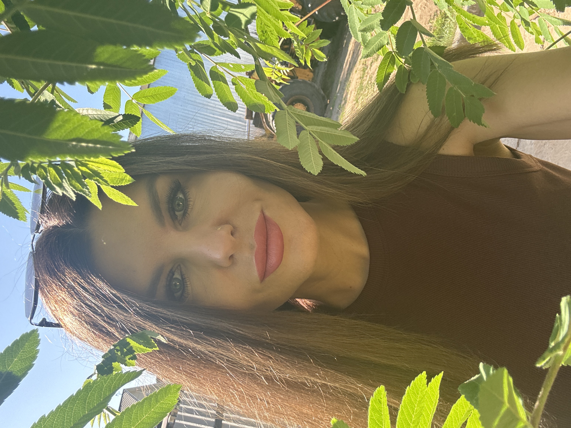 A woman is posing for a selfie, with green foliage framing her face. She has light-colored eyes, light skin, and long brown hair. She is wearing a brown top and lipstick. A pair of sunglasses rests on her head. In the background, there is a blurred glimpse of what appears to be a tractor or other agricultural machinery and a corrugated metal structure, suggesting an outdoor, possibly rural or agricultural setting. The photograph was likely taken outdoors in natural light.
Although the photograph includes a tractor in the background, it is not possible to determine the location as Dubovaya, Russia based on the visual information provided.