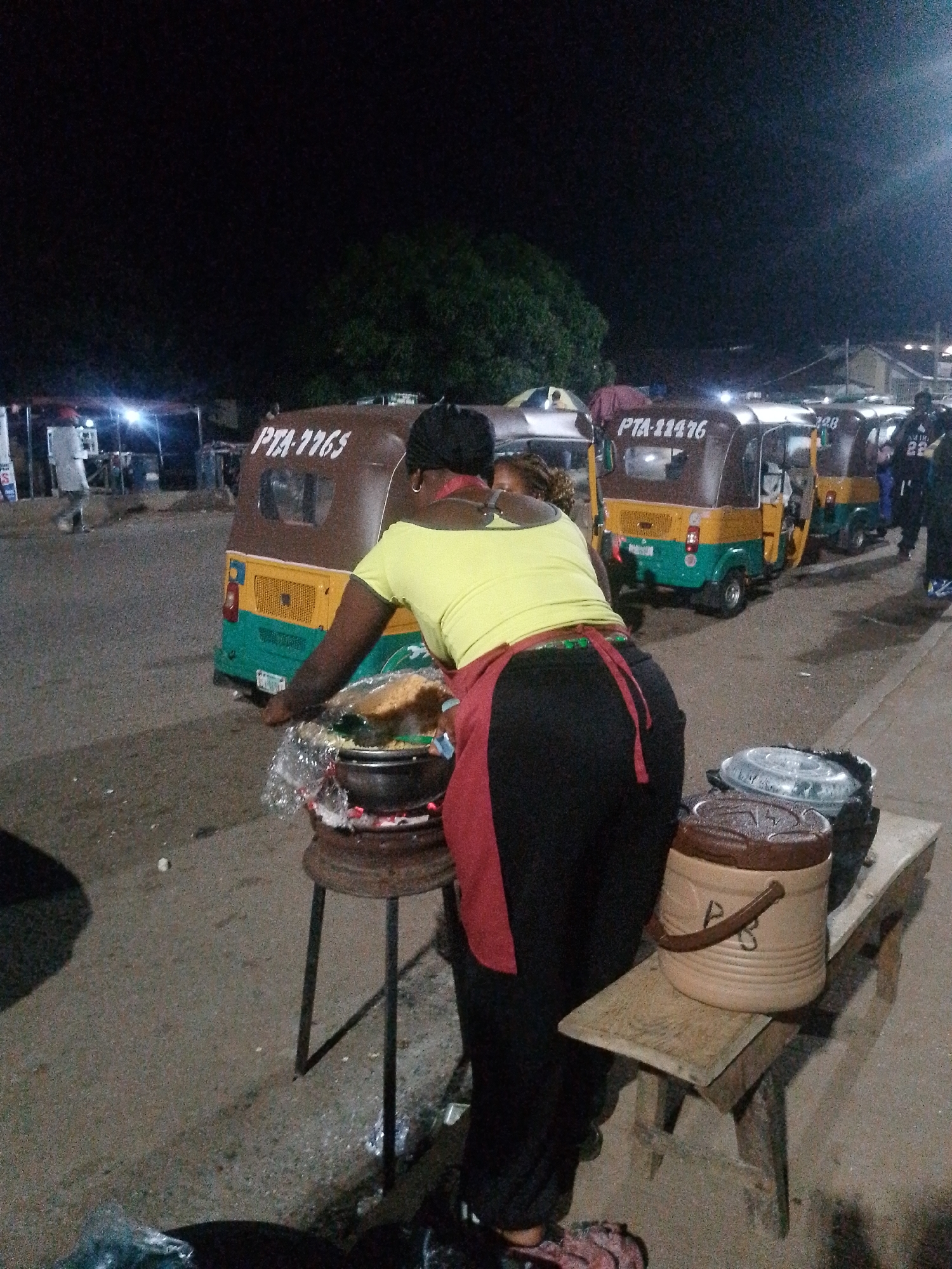 This is a night-time photograph taken in Bukuru, Nigeria. The scene depicts a street vendor, a woman wearing a yellow shirt and a red apron, cooking over an open fire. She is standing beside a pot that is covered with plastic wrap and is placed on a metal stand with a fire underneath. In the background, several auto-rickshaws, also known as 'Keke Napep', are parked, suggesting a transportation hub or a busy street. One of the Keke Napep vehicles has the license plate 'PTA-7765' visible, and another has 'PTA-11476'. The ground is paved with asphalt. A wooden bench with containers on it is visible in the foreground. The overall lighting suggests artificial streetlights and the glow from the cooking fire.
