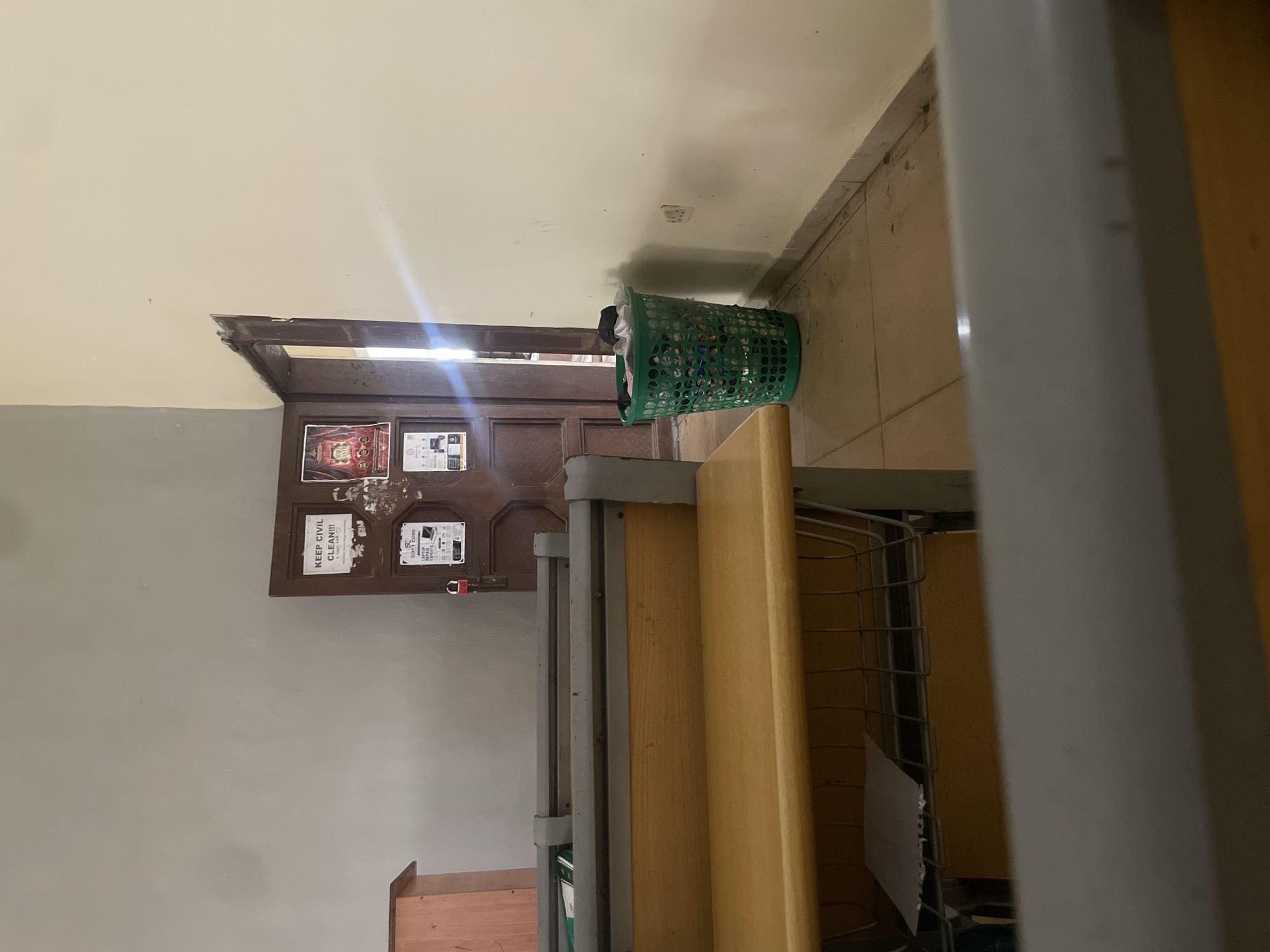 The image depicts an interior scene, likely a classroom or office space, in Ihiagwa, Nigeria. In the foreground, the edge of a wooden desk with a metal frame is visible. Behind it, a doorway leads to another room, with a dark wooden door bearing several posted notices. One notice reads "KEEP CIVIL CLEAN!!!" with some illegible text and drawings underneath. Another notice displays the "SC SOFT CORE" logo and advertises "LAPTOP SALES & REPAIRS EXPERTS." A red padlock is attached to the door. To the right of the doorway, a green plastic laundry basket is overflowing with clothes. The room appears sparsely furnished with tiled flooring and light-colored walls. A wooden cabinet is partially visible on the left side of the frame. The overall impression is one of a functional but somewhat cluttered space.