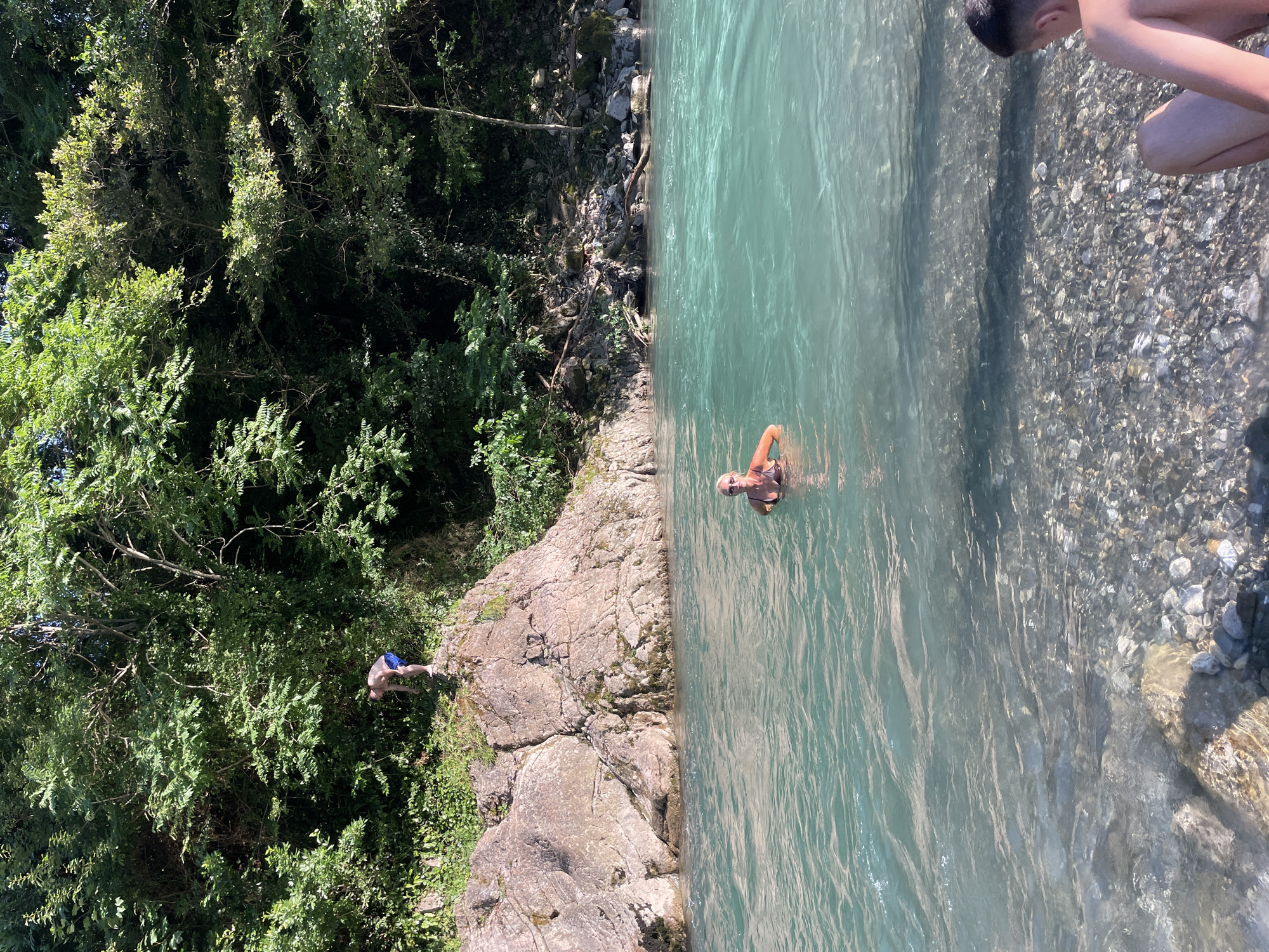 The image shows a scenic view of a river with lush green trees and rocky terrain on the left, and clear, turquoise water on the right. Two individuals are visible. One person is in the water, near the shore, partially submerged and looking towards the camera. Another person is standing on a rocky ledge on the left side of the image, facing away from the camera and appearing to be preparing to jump into the water. The sun is shining, casting dappled light on the water and foliage, suggesting a bright, sunny day. The overall atmosphere appears relaxed and recreational, indicative of a summer day spent enjoying nature and the water. The location appears to be a natural outdoor setting, possibly a river gorge or a secluded swimming spot. No visible text is present in the image.