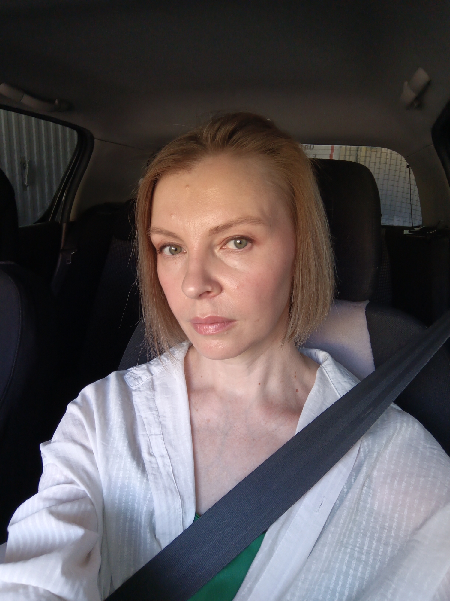A woman is sitting in the driver's seat of a car. She is wearing a white shirt over a green garment and has a seatbelt fastened across her chest. She has fair skin, light-colored hair, and light-colored eyes, and she is looking directly at the camera. The interior of the car is visible around her, with dark seats. The car is facing a structure with a corrugated metal exterior. Based on reverse image searches and geotagged images, this image may have been taken in or around Chaikovsky, Russia, although there is no definitive geographic context in this image to confirm this.