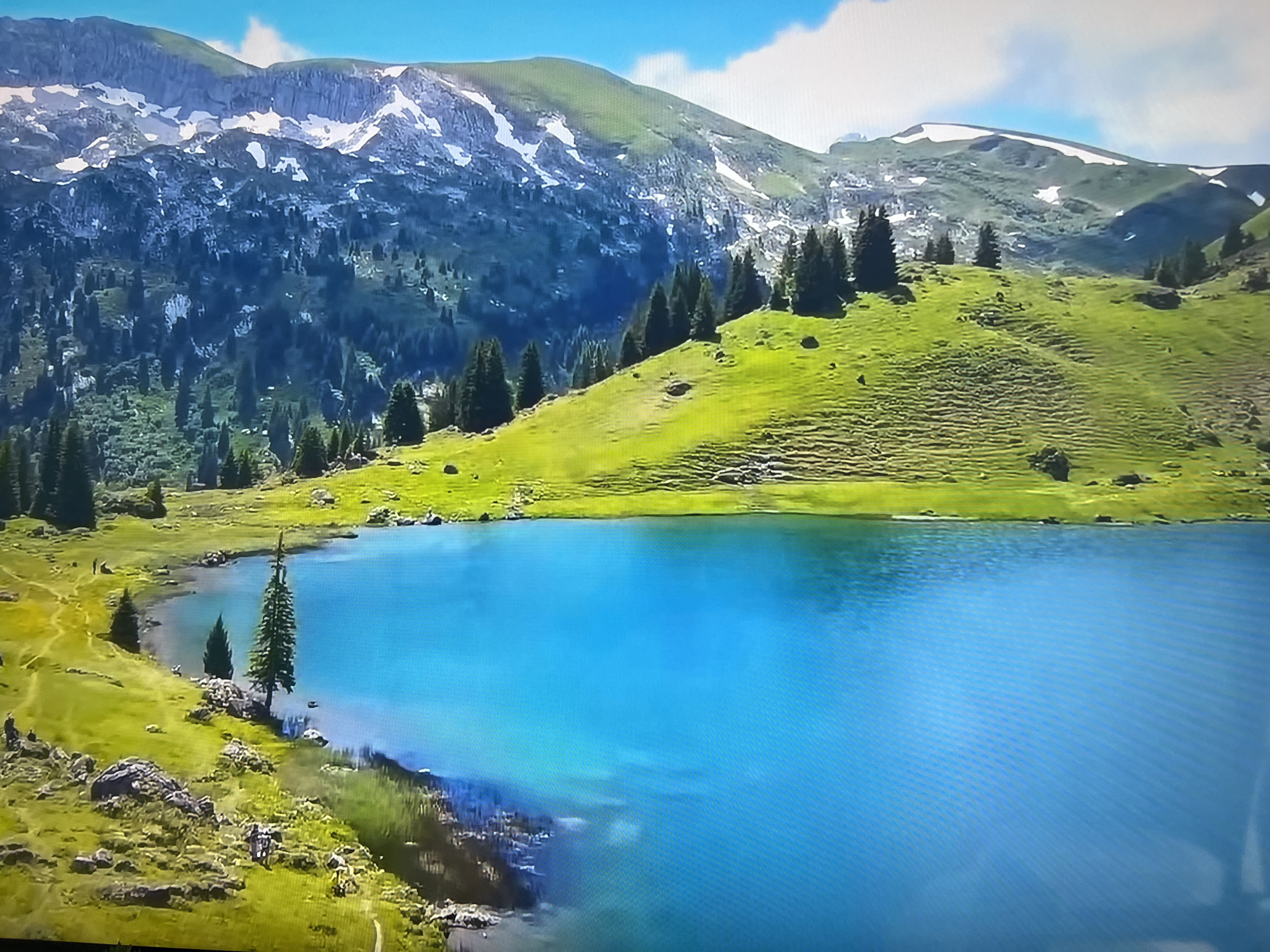 The image shows a mountain landscape with a blue lake in the foreground. The mountains are green with patches of snow. There are scattered pine trees on the hillside. The sky is blue with some clouds. The location is not in Basingstoke and Deane, United Kingdom.