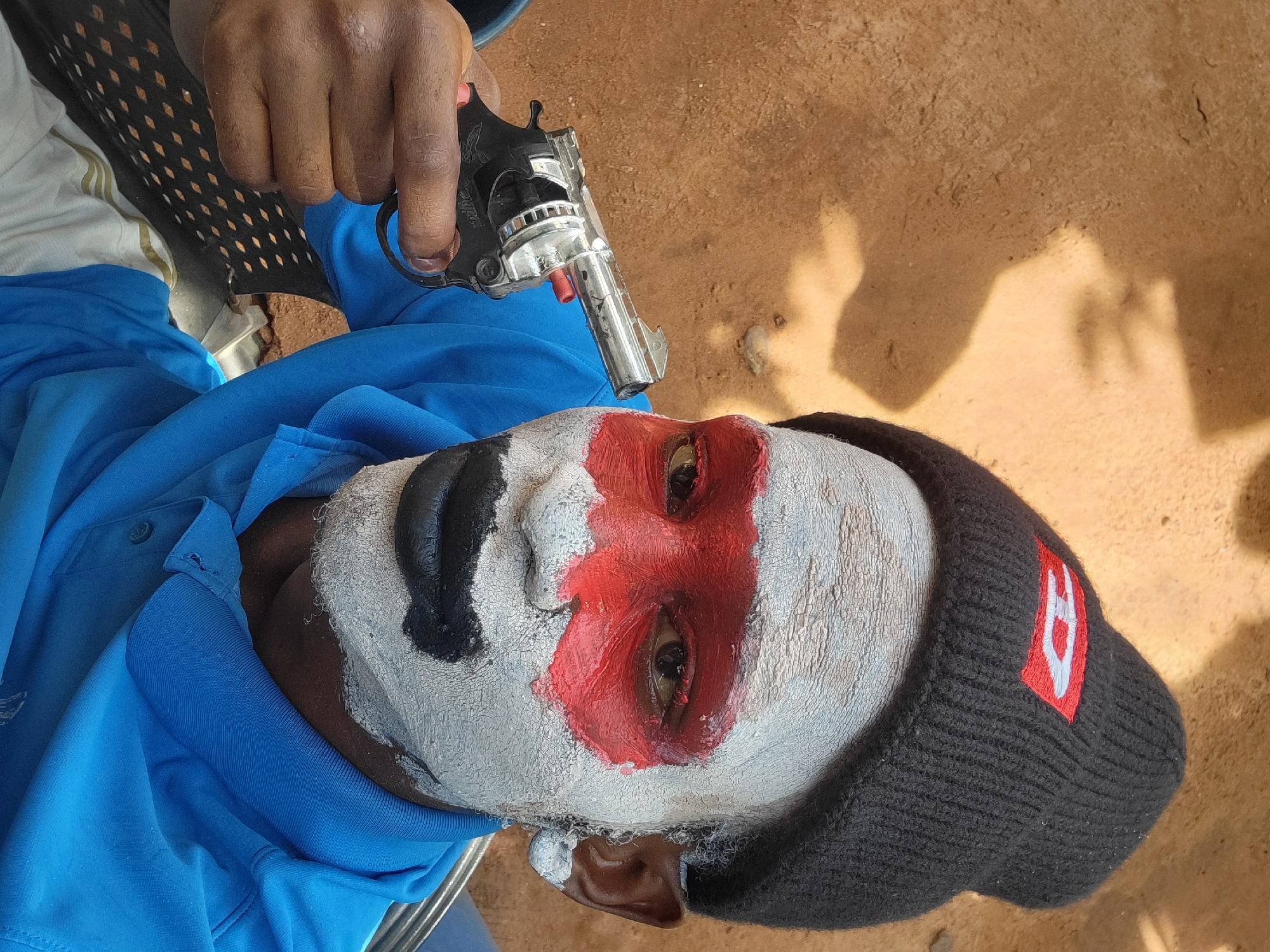 A person lies supine, wearing white and red paint around the eyes and forehead, and black paint on the left side of the face. They wear a black knit cap with a red and white emblem on the side and a blue shirt. A hand holding a silver and black handgun points toward the person's head. The setting appears to be outdoors, possibly in Nkpor, Nigeria, based on visual characteristics. The surrounding ground is dirt-colored. The image depicts a mock execution.