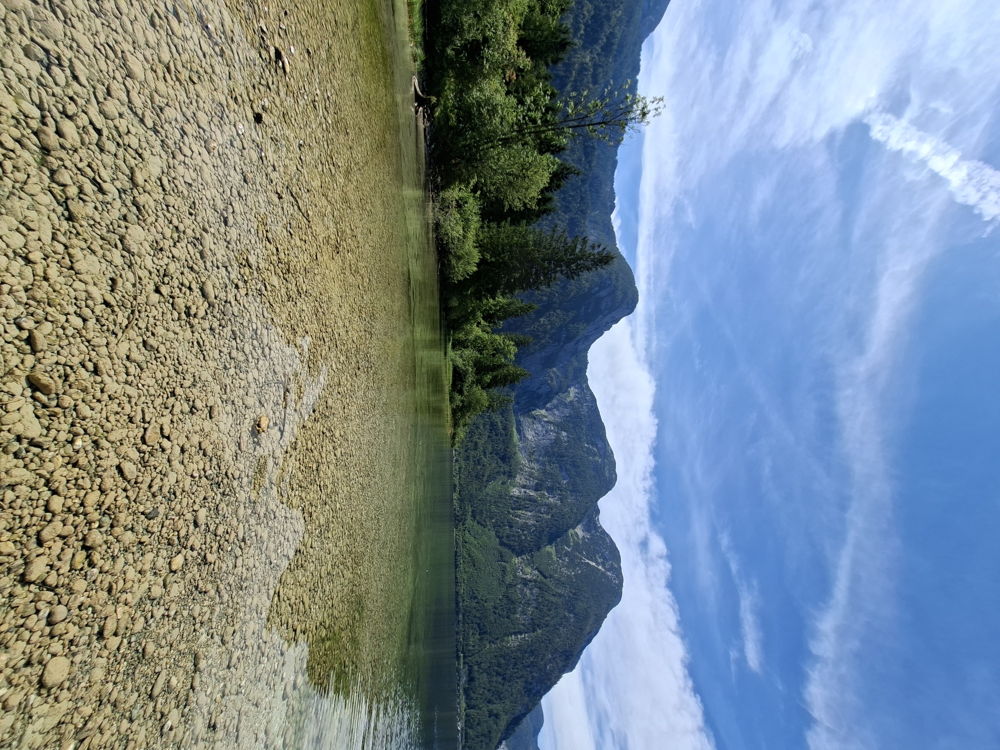 The image shows a clear, shallow lake with a rocky bottom in Obertraun, Austria, at the southern tip of Hallstätter See. The water is so transparent that the pebbles and stones on the lakebed are clearly visible. In the background, lush green trees and vegetation cover the lower slopes of imposing, mist-shrouded mountains. The sky above is a brilliant blue, dotted with wispy white clouds, suggesting a pleasant day. The scene evokes a sense of natural tranquility and the pristine beauty of the Austrian Alps. There are no people or animals visible in the image. The composition is dominated by the natural landscape.