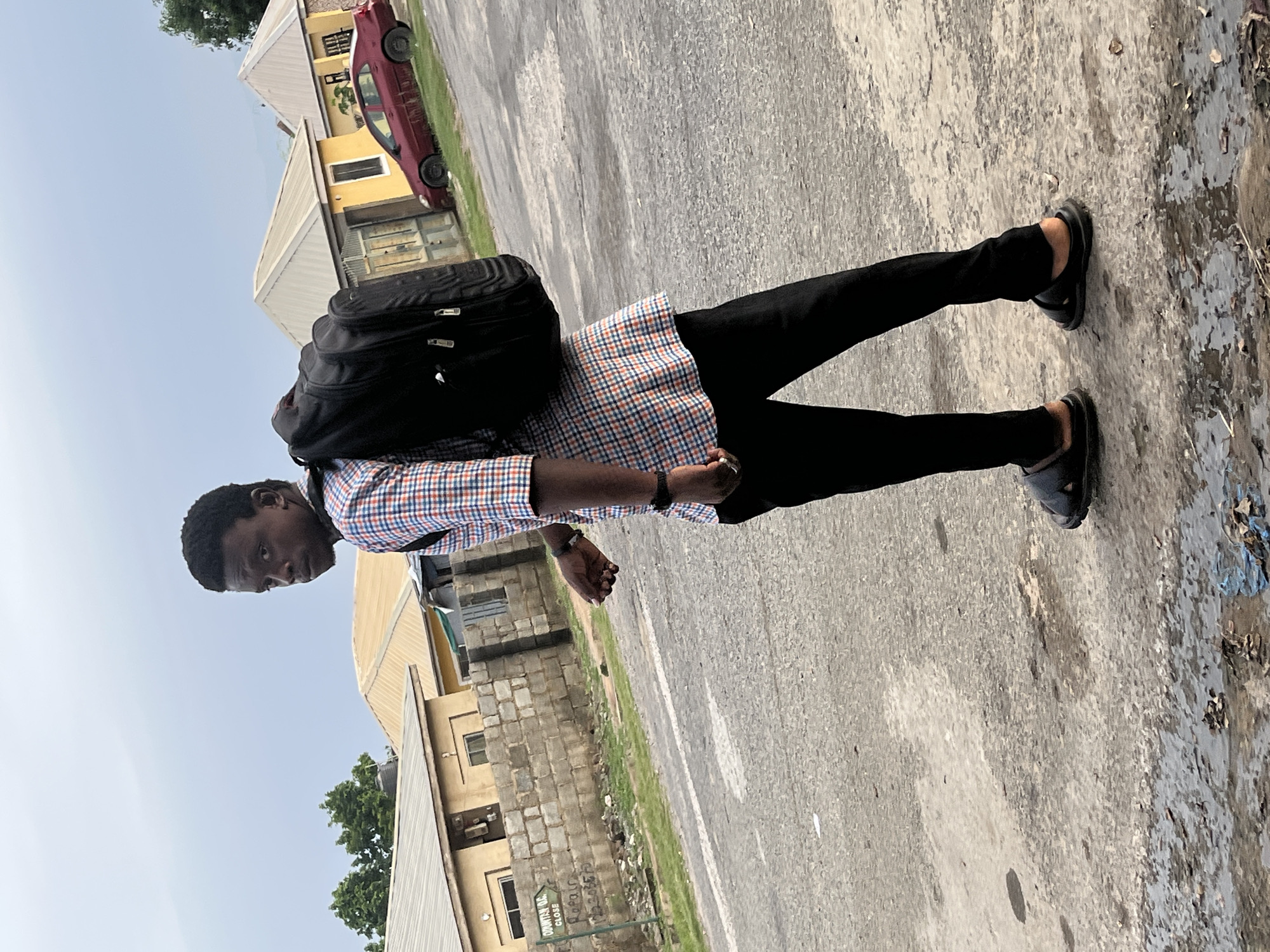 A young Black male wearing a plaid shirt, black pants, and sandals walks on a paved street in Abuja, Nigeria. He is carrying a black backpack. In the background, there are buildings with yellow and white roofs, a red car, and a brick wall with a sign. The sky is overcast.