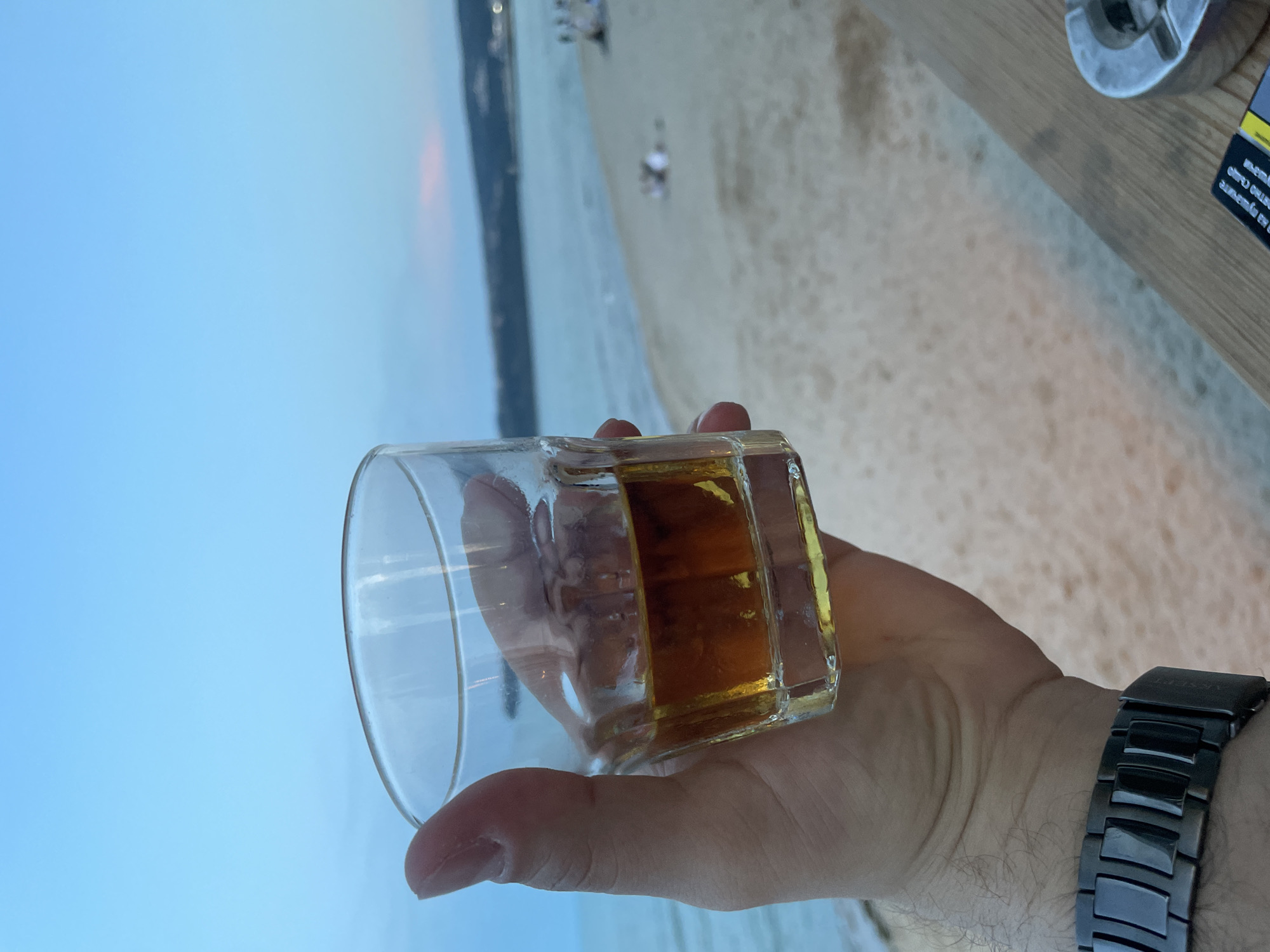 A first-person view depicts an adult male's hand holding a clear, ridged glass tumbler containing an amber-colored liquid, likely an alcoholic spirit, at dusk on the Black Sea coast in Varna, Bulgaria. The hand, wearing a dark metallic link watch on the wrist, grips the glass which is approximately one-third full. In the lower right foreground, a light-colored wooden surface supports a partial view of a metallic ashtray and a dark-colored cigarette pack featuring Cyrillic text, including "А на пушачите" (And to smokers).
The background reveals a wide, light-sandy beach extending towards calm, light-blue seawater. Several blurred human figures are visible on the distant beach, engaged in leisure activities. Beyond the sea, a dark, undulating landmass, consistent with coastal hills or cliffs, appears on the horizon with scattered faint lights. The sky overhead is a clear, gradient light blue, subtly transitioning to a faint pinkish hue on the right horizon, indicating twilight or early evening.