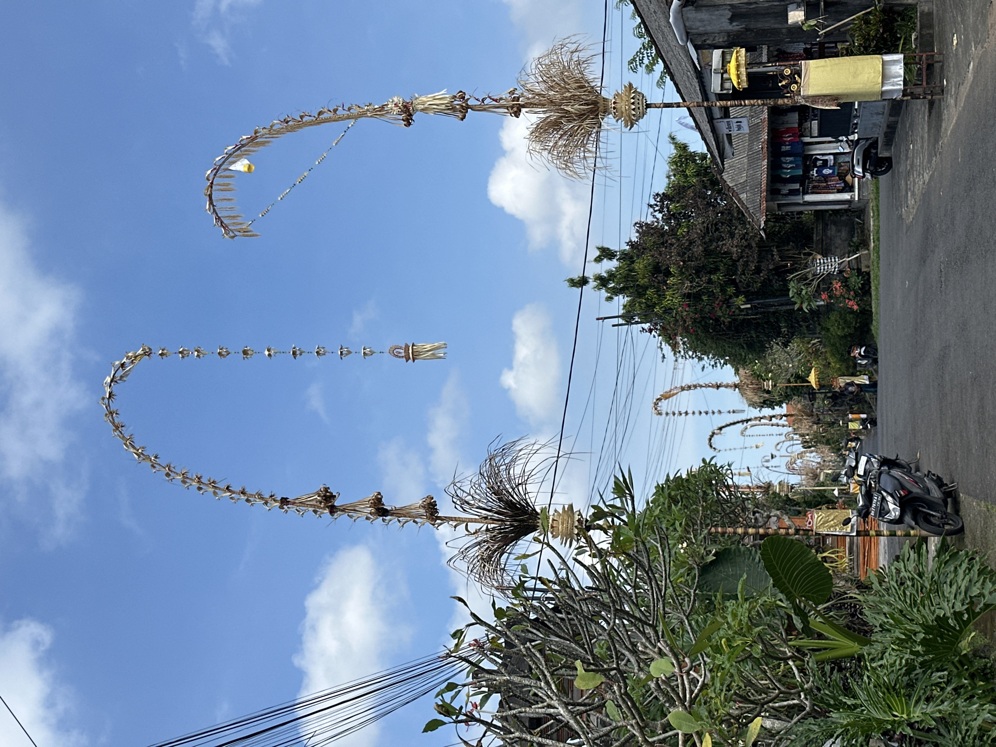 In the city of Tampaksiring, Indonesia, a street is adorned with tall, arched decorations made of natural materials, possibly for a festival. The decorations curve over the road, with smaller ornaments hanging from them. Along the sides of the street, there are lush green plants and trees, as well as buildings that appear to be shops, with items displayed outside. Several motorcycles are parked on the roadside. The sky is blue with scattered white clouds. The scene suggests a vibrant cultural celebration or event taking place in the town.