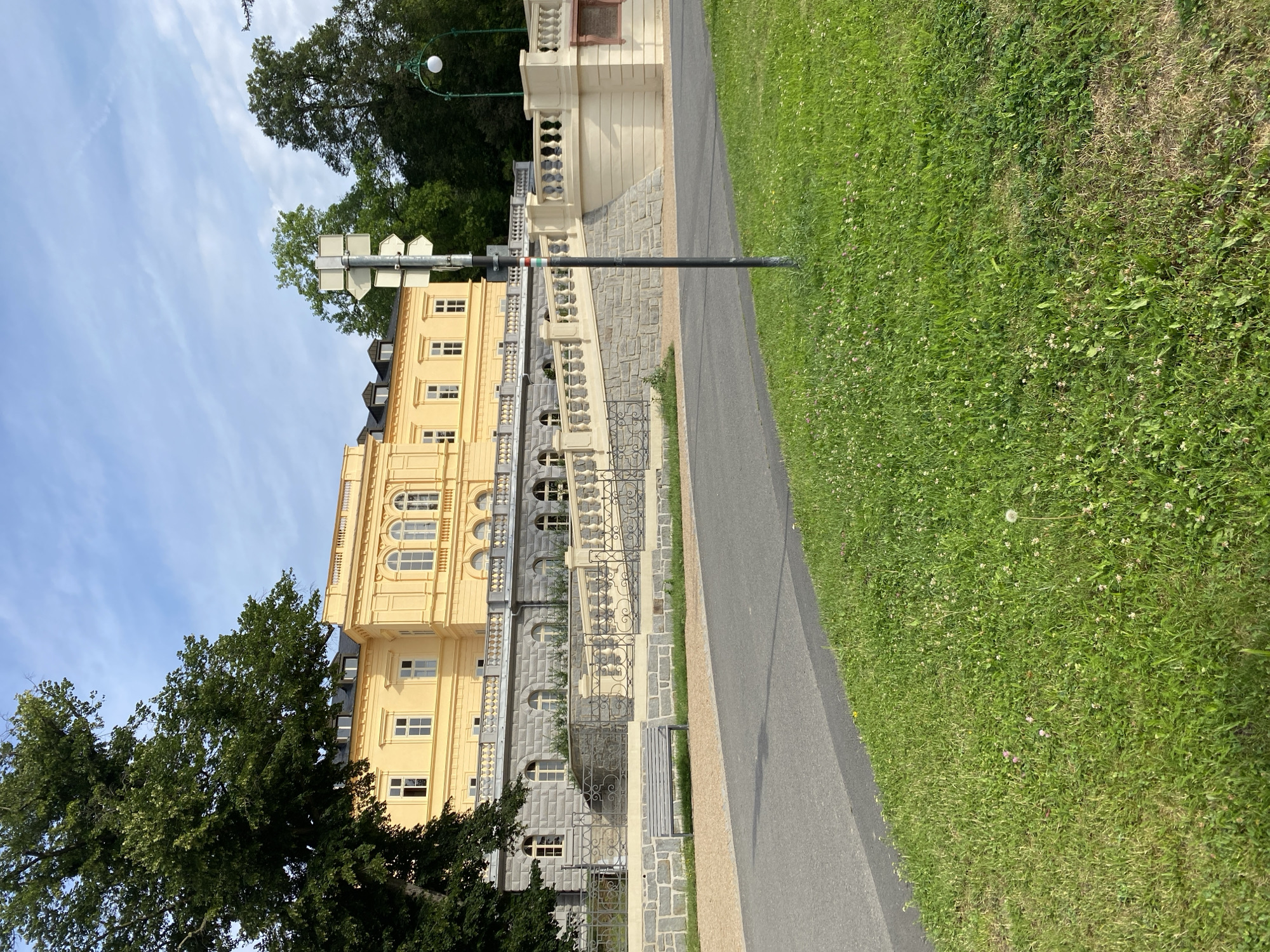The image shows a yellow classical building with a gray stone lower facade, located in Bílina, Czechia. The building has arched windows on the lower level and rectangular windows on the upper level, topped with a balustrade. A wide stone staircase with balusters leads up to the building. In the foreground, a paved path runs alongside a grassy area with scattered wildflowers and dandelions. A metal bench sits on a gravel path adjacent to the staircase. A lamppost with a spherical light fixture is visible to the right of the building. A signpost with multiple arrow indicators stands near the lamppost. Trees with dense green foliage surround the building and a large tree is on the left side of the image. The sky above is blue with scattered white clouds.