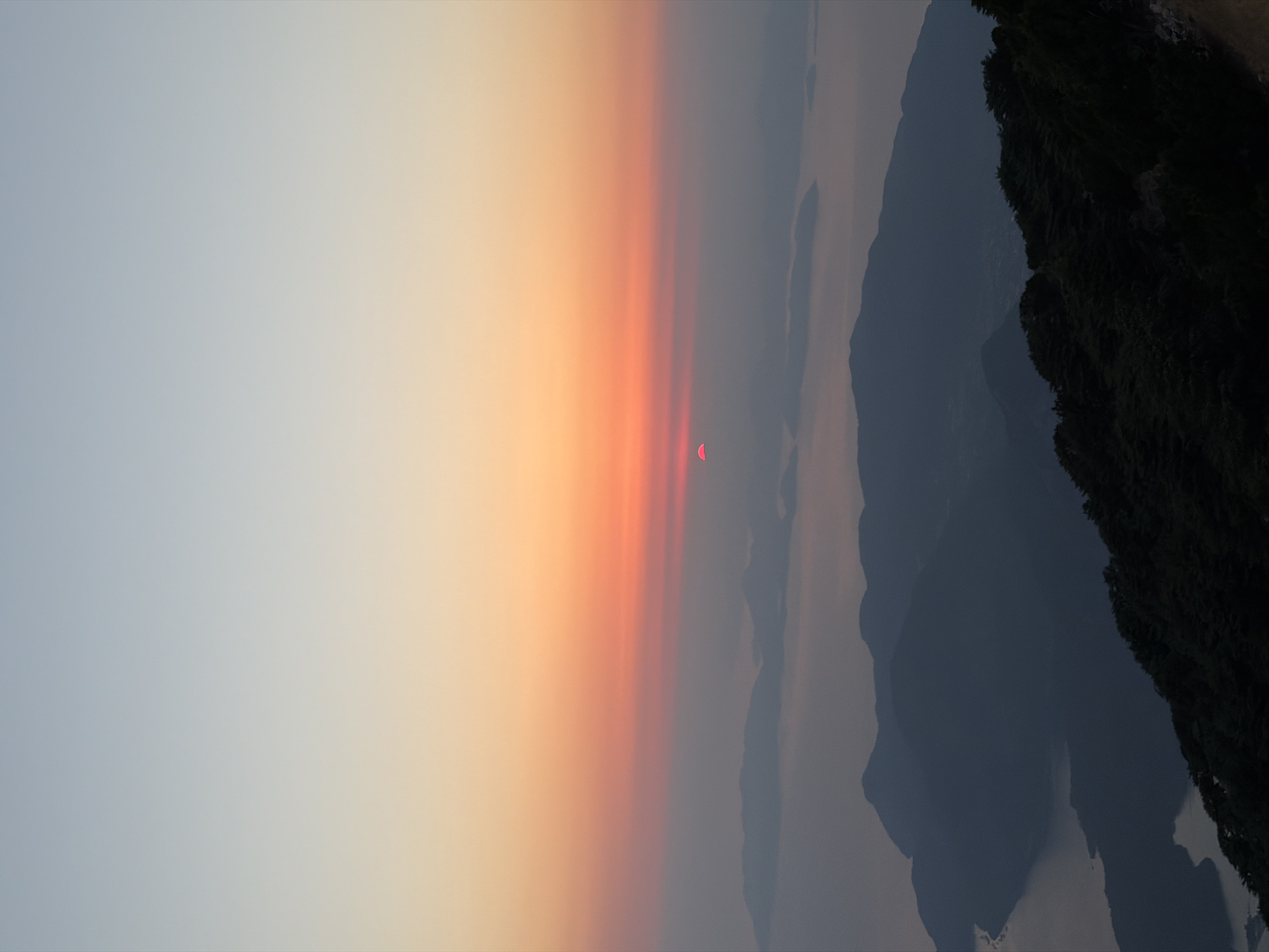 An elevated, vertical, full shot captures a sunrise over a hazy landscape of islands and mountains. The sky transitions from a pale blue at the top to an intense orange and red hue near the horizon, where the sun, a small, vibrant red semi-circle, is visible just above the distant islands. These islands, shrouded in mist, are silhouetted against the glowing sky, appearing as dark, rounded shapes. In the foreground, a range of dark, rugged mountains, densely covered in green foliage, slopes down from the bottom right of the frame, leading the eye toward the sea and islands. The scene suggests a natural, serene environment at dawn. The location of Fethiye, Turkey is not discernible from the visual elements alone, as the image depicts a general seascape with mountainous terrain and islands. There are no people or specific man-made objects identifiable in the image.