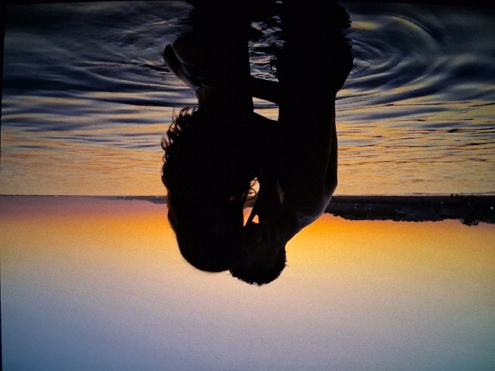 The image depicts two individuals in full silhouette, an apparent male and female, embracing and kissing at an undetermined waterfront location during either sunrise or sunset. The male figure is positioned on the left, shirtless, leaning into the female figure on the right, who has long, curly hair. Their faces are pressed together, and their bodies are in close contact. The background features a sky transitioning from light blue at the top to vibrant orange and yellow hues at the horizon, indicative of dusk or dawn. A faint, dark line along the horizon suggests distant land or structures. To the right, a body of water shows ripples and reflects the sky's colors in tones of dark blue, orange, and yellow. The specific location in the city of Unknown, Unknown, cannot be determined beyond being an outdoor waterfront setting.
