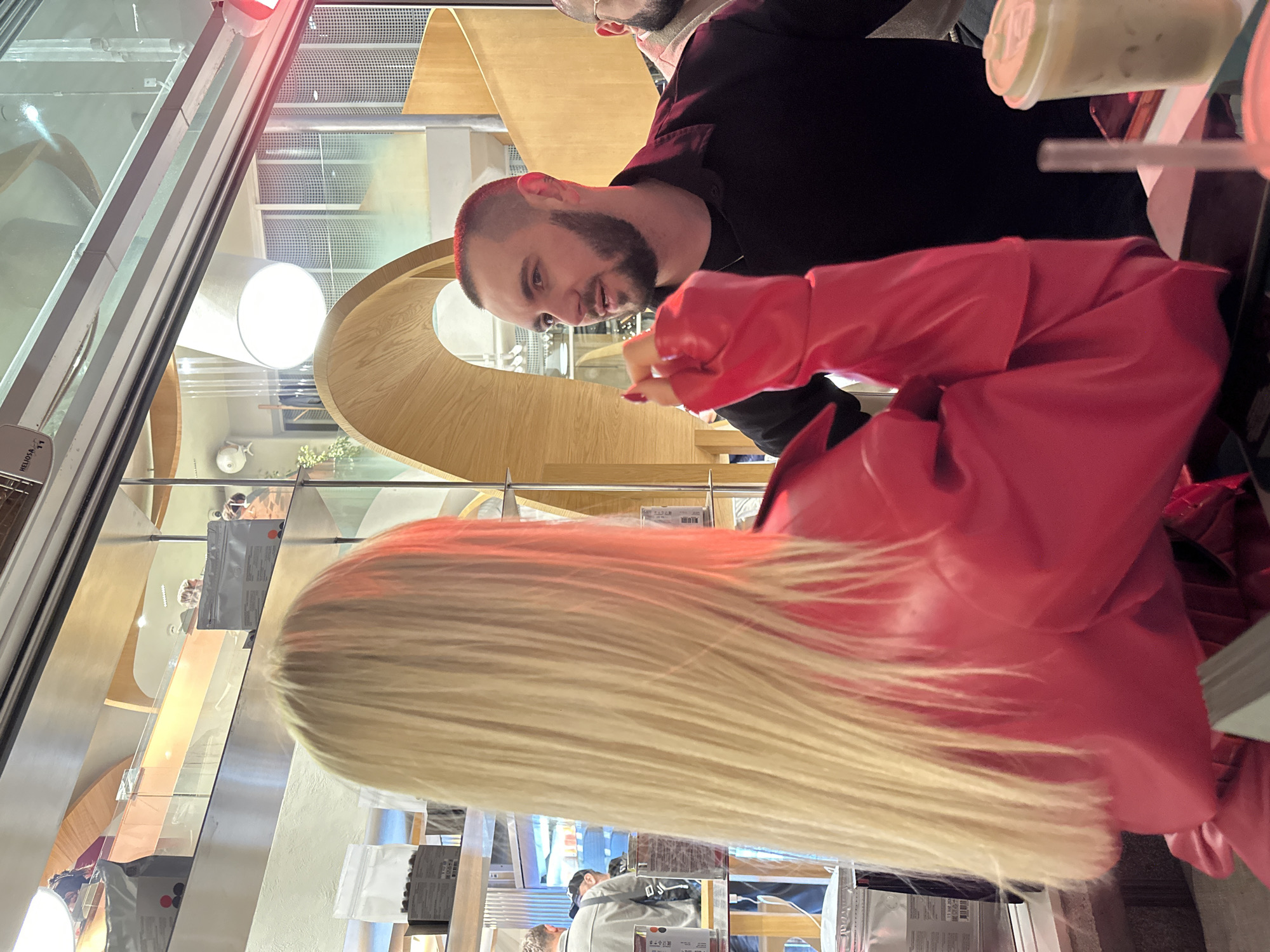 A man with a short beard and dark hair in a black sweater smiles at a woman with long, blonde hair wearing a red leather jacket. They are inside a building with a modern interior featuring wooden arched structures. Transparent packaging is visible on a shelf beneath a glass partition. A city of Yerevan, Armenia location is suggested by the image's content.