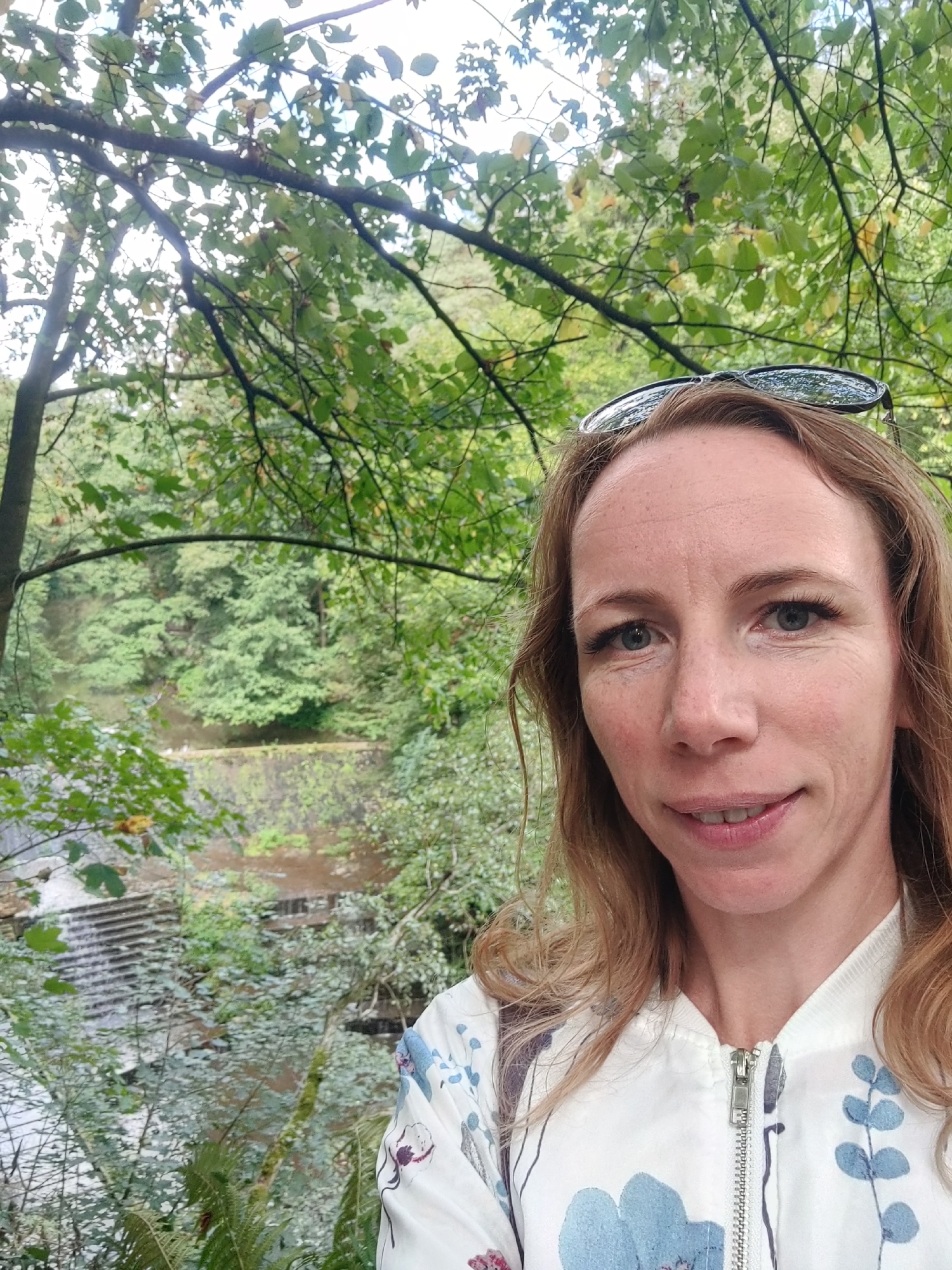 A woman with blonde hair and blue eyes smiles at the camera in front of a wooded area. She is wearing a white bomber jacket with a floral pattern and sunglasses are perched on her head. In the background, a waterfall cascades down a rocky embankment into a body of water. The scene appears to be a natural setting, possibly a park or hiking trail. This location is likely to be the famous Liebethaler Grund in Lohmen, Germany, known for its scenic beauty and waterfalls.