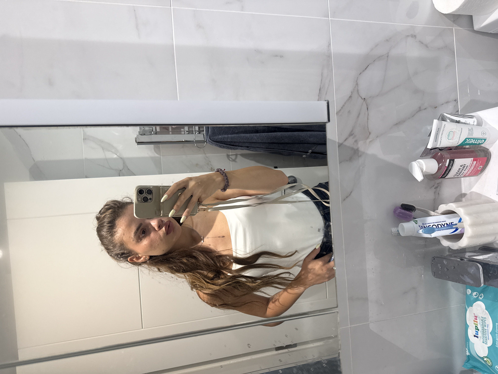 A woman is taking a selfie in a bathroom mirror. She has long, brown, wavy hair and is wearing a white tank top and dark jeans. Her left hand is in her pocket, and she is holding her phone in her right hand. In the background, the bathroom is modern with marble tile. Visible on the counter are a toothbrush holder with toothbrushes and toothpaste, a bottle of Neutrogena face wash, another tube of toothpaste, and a package of baby wipes. There are no specific details in the image to indicate the location is Throop, United Kingdom.