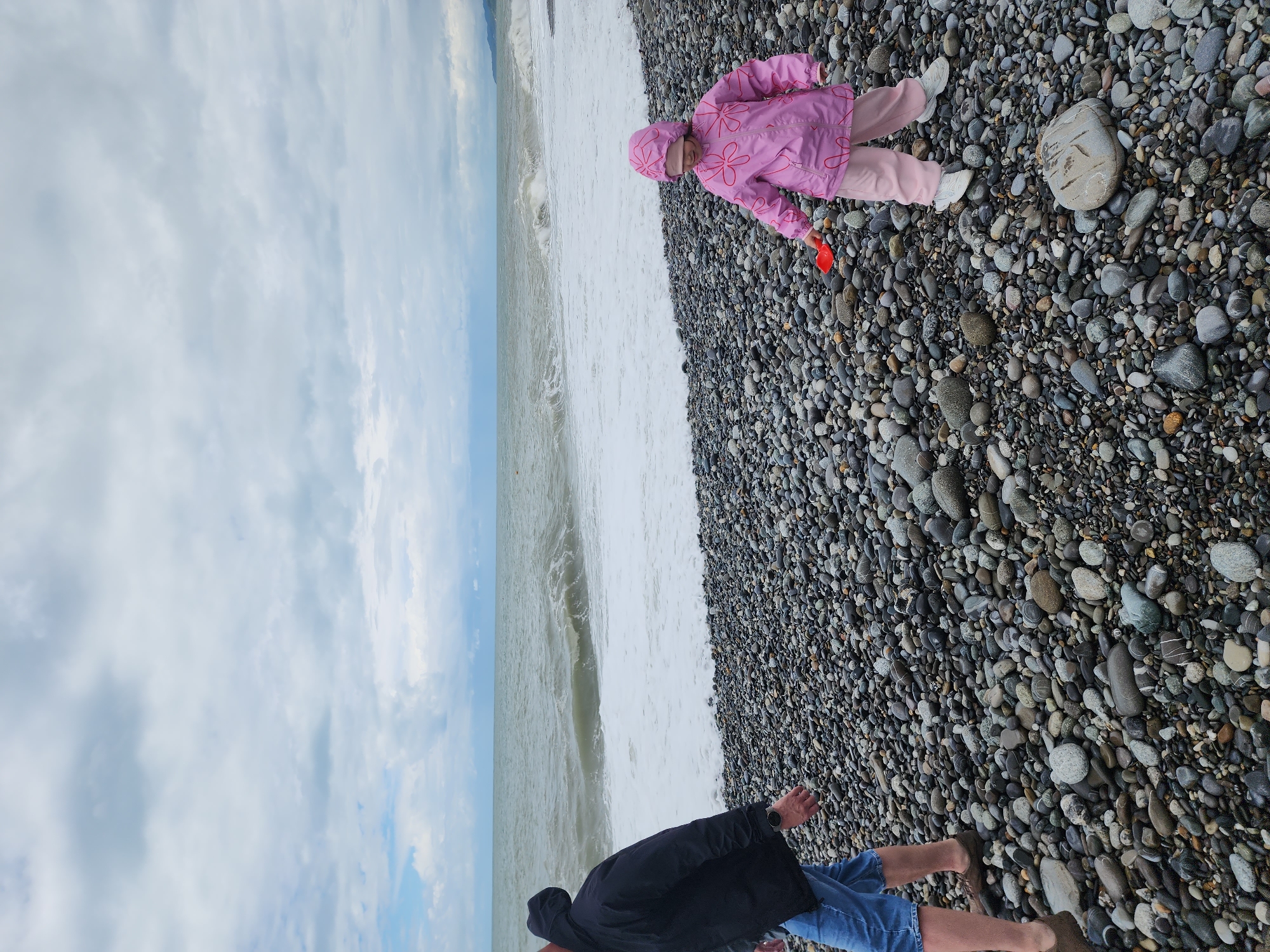 The image depicts a pebble beach in Sochi, Russia. A child in a pink floral jacket and matching hat sits on the pebbles near the shoreline, holding a small red shovel. An adult, wearing a dark jacket and blue shorts, stands further down the beach, facing the ocean. White waves are visible at the edge of the beach, and the sky is overcast with cloudy skies.
