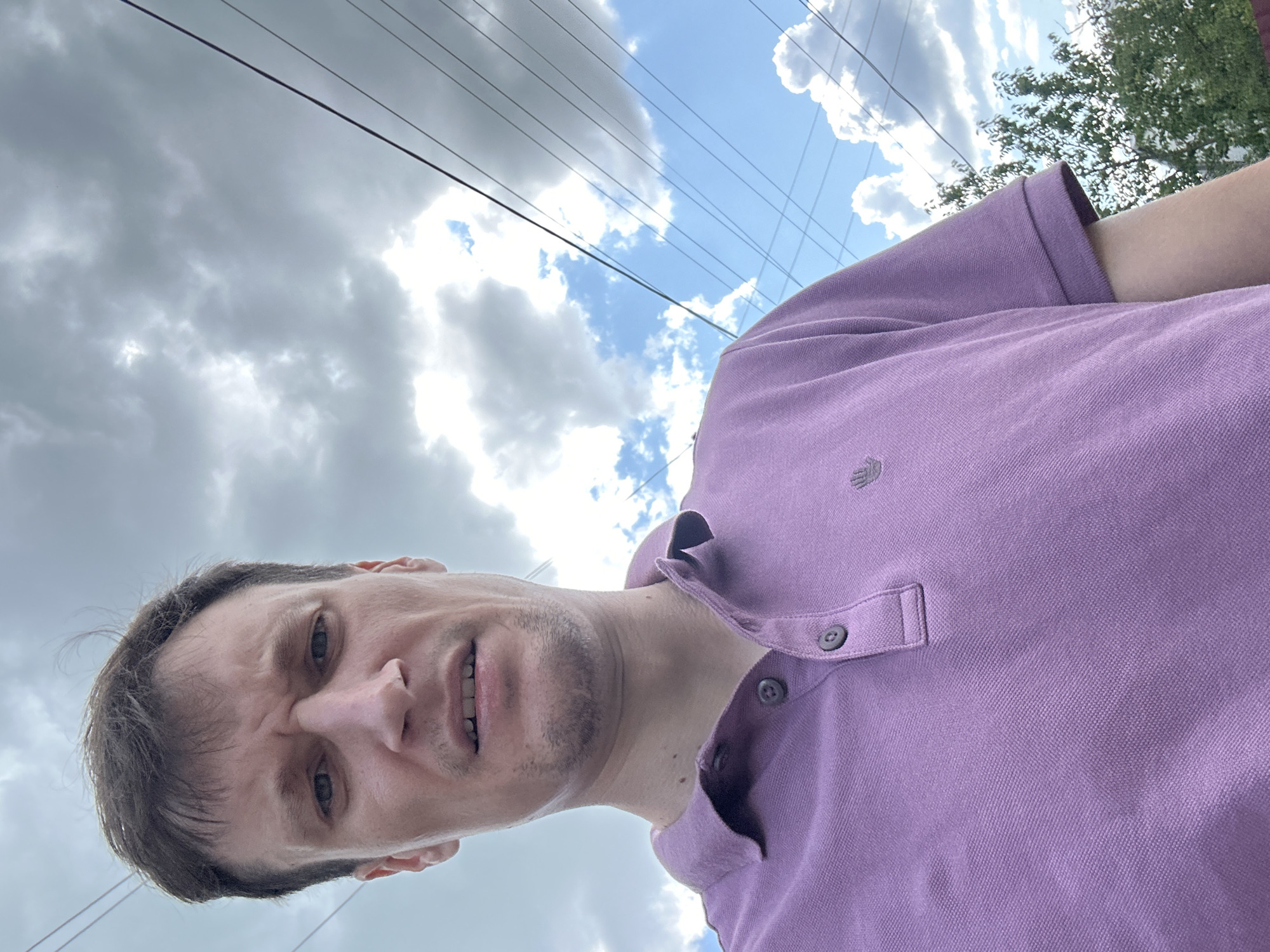 A man is taking a selfie outdoors in Makiv, Ukraine, with a cloudy sky and overhead power lines visible. He is wearing a purple polo shirt and has a concerned expression. Trees and foliage are in the background.