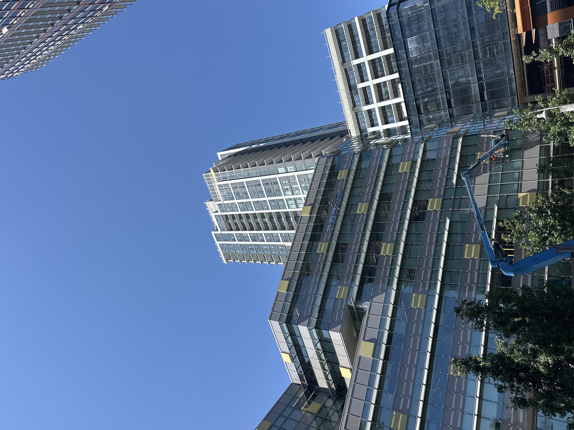 In the city of New Westminster, Canada, on a bright, clear day, the image captures an upward view of modern architecture against a vibrant blue sky. The composition features several multi-story buildings, including a tall, slender residential tower with numerous windows and balconies, positioned behind a lower, angular office building characterized by a facade of dark glass and grey paneling, accented with yellow square motifs. Another smaller office building with white framing and dark glass is visible to the right of the main structure. In the foreground, a blue boom lift is positioned, with two individuals working in the elevated basket, attending to the exterior of the office building. Green trees are visible at the base of the buildings, suggesting an urban park or streetscape. The scene conveys a sense of ongoing maintenance or construction activity within a contemporary urban environment.
