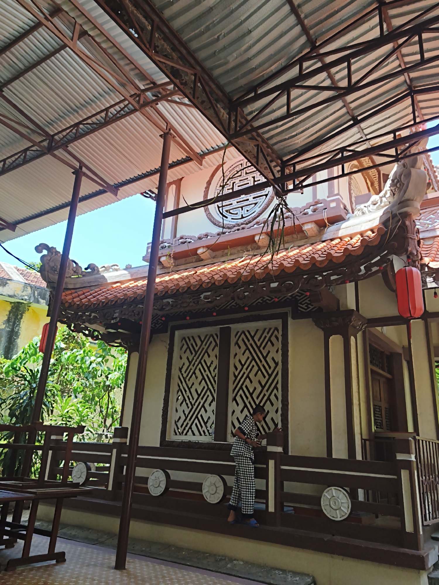 The image depicts an outdoor scene at a Vietnamese pagoda in Nha Trang, Vietnam. A person dressed in a black and white patterned outfit and blue sandals stands near a railing, facing away from the camera and appearing to be looking at something on the railing. The pagoda features traditional architecture with a tiled roof and ornate details. A red lantern hangs from the eaves. In the foreground, a wooden bench or table is visible. The surrounding area is lush with green foliage. The sky above is a clear blue. The scene is bathed in natural daylight.