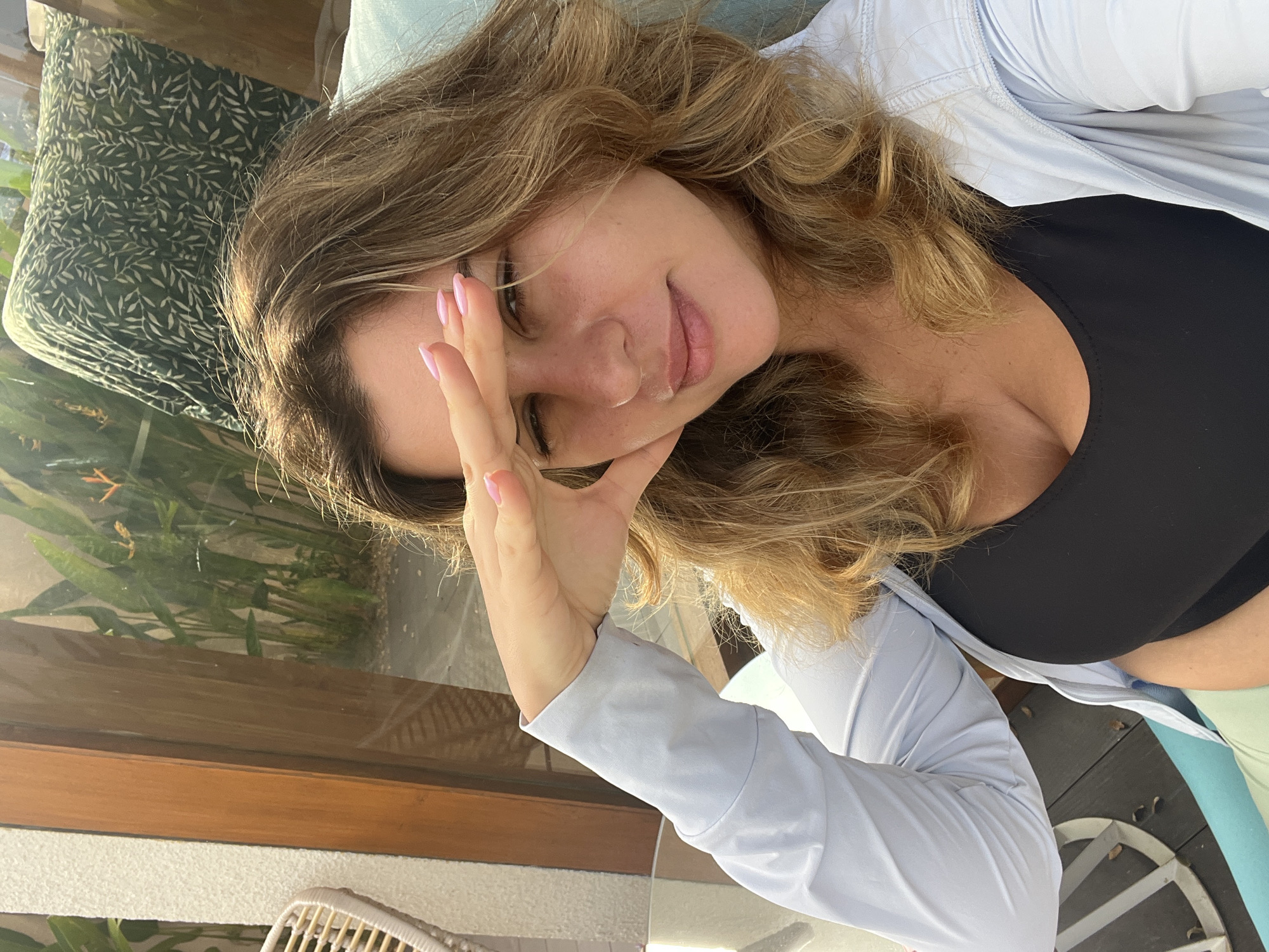 A woman is captured in a selfie taken outdoors, possibly on a terrace or balcony. She is wearing a black tank top and a light blue, open zip-up jacket. Her right hand is raised to shield her eyes from the sun, with her fingernails painted pink. She has wavy, blonde-brown hair and fair skin. In the background, lush green plants and foliage are visible, suggesting a tropical or garden setting. The overall lighting indicates it is daytime, with sunlight filtering through. The scene does not contain any elements that place it specifically in Kediri, Indonesia.