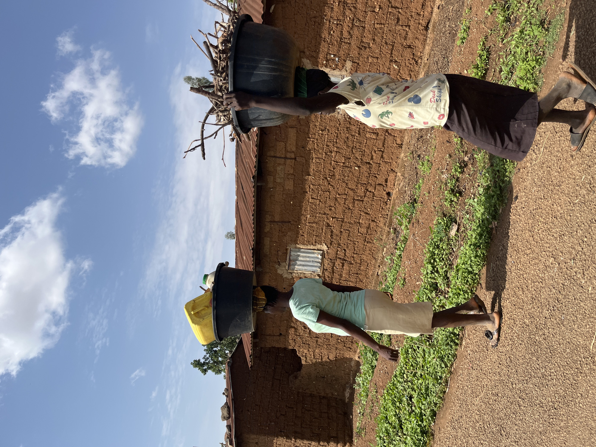 Two women are carrying items on their heads while walking on a dirt road in Barakin Momo Dogo, Nigeria. The woman on the left is carrying a black bucket with a yellow container and a white plastic bottle on top of it. The woman on the right is carrying a black bucket filled with sticks. Both women are walking past buildings made of mud bricks. The sky is blue with some clouds. The ground is covered with dirt and some patches of green plants.