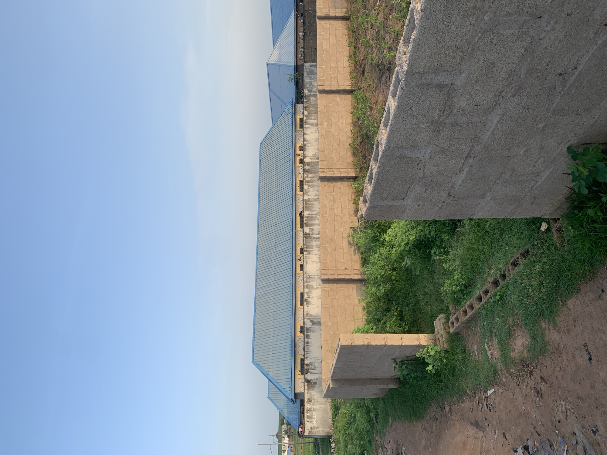 The image depicts an outdoor scene in the city of Jalingo, Nigeria, focusing on a construction site. In the background, a series of buildings with light blue, corrugated metal roofs are visible, separated by a low concrete wall topped with barbed wire. These buildings appear to be single-story structures with yellow decorative bands running below the roofline and windows along their facades. In the foreground, several low walls constructed from concrete blocks divide the area. One wall, closer to the viewer, is prominently featured on the right, made of large, rough-textured concrete blocks. Another wall, further back and to the left, is also made of concrete blocks and appears to be part of the perimeter of the construction site. The ground is a mixture of dirt paths, overgrown green grass, and areas of tilled earth where young crops are growing. The sky is a clear, light blue, indicating daylight. The overall impression is one of ongoing construction and development in a semi-urban or rural setting within Jalingo.