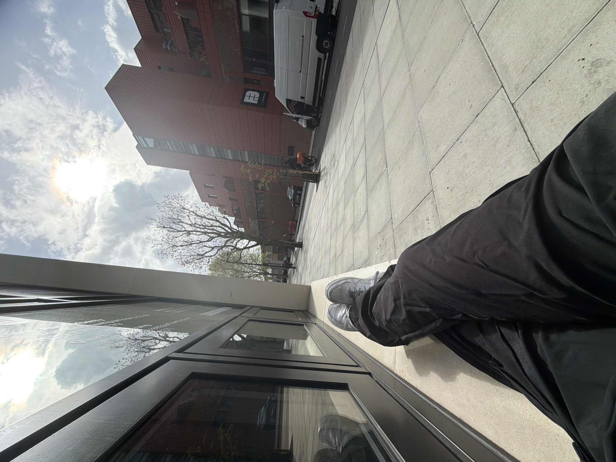 A low-angle urban street scene shows a person's lower legs and feet, clad in black trousers and grey sneakers, resting on a beige ledge or low wall in the foreground. The person is positioned beside a modern building with large, dark-framed windows that reflect the bright, partly cloudy sky and surrounding environment.
Across the street, a tall, reddish-brown brick building with a distinctive stepped design stands prominently. On its facade, a white, stylized logo, possibly "HB" or "HE," is visible, with the words "FITNESS & WELLBEING" faintly discernible below it. Parked vehicles line the street in front of this building, including a white van, an orange scooter, and a dark car. Trees with bare branches suggest either late winter or early spring. The ground is paved with light-colored flagstones.
The sky in the upper left is bright and partly cloudy, with the sun casting a strong glare and exhibiting a slight rainbow halo effect. The bright daylight indicates it is likely midday or early afternoon. Reflections on the window next to the person show faint, illegible white text, possibly website addresses ending in ".com" or ".co.uk". The overall scene depicts a calm urban environment, consistent with a street in London, United Kingdom.