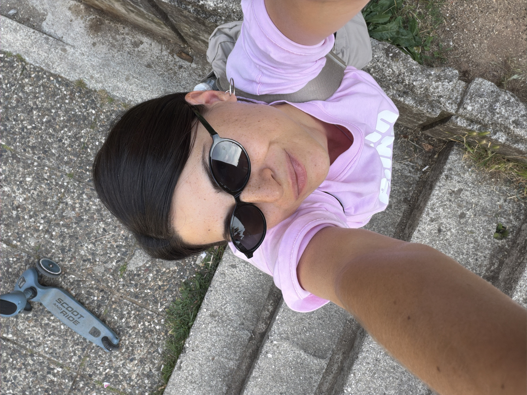 A person takes a selfie while standing on concrete steps. The person wears sunglasses and a light purple shirt. To the left of the person, on the ground, is a light blue scooter with the words "SCOOT AND RIDE" printed on it. The location is not specified within the image, and cannot be confirmed.