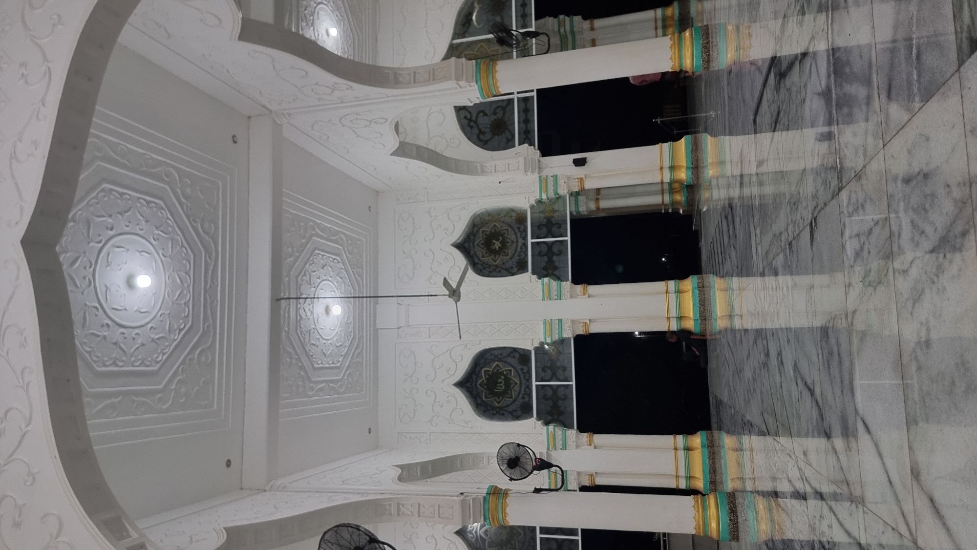 The image shows the interior of a mosque with white ornate ceilings and pillars. The floor is polished marble, reflecting the pillars and the ceiling lights. There are black archways leading into darker areas, and in the foreground, a ceiling fan is visible. On the wall, above the archways, are decorative Islamic calligraphy panels. Two black electric fans are mounted on white pillars. A person is seated in the darkness beyond one of the archways on the left, and another person is visible in the distance on the right. The overall impression is one of grandeur and religious solemnity. The location is identified as Lapang Timu, Indonesia.