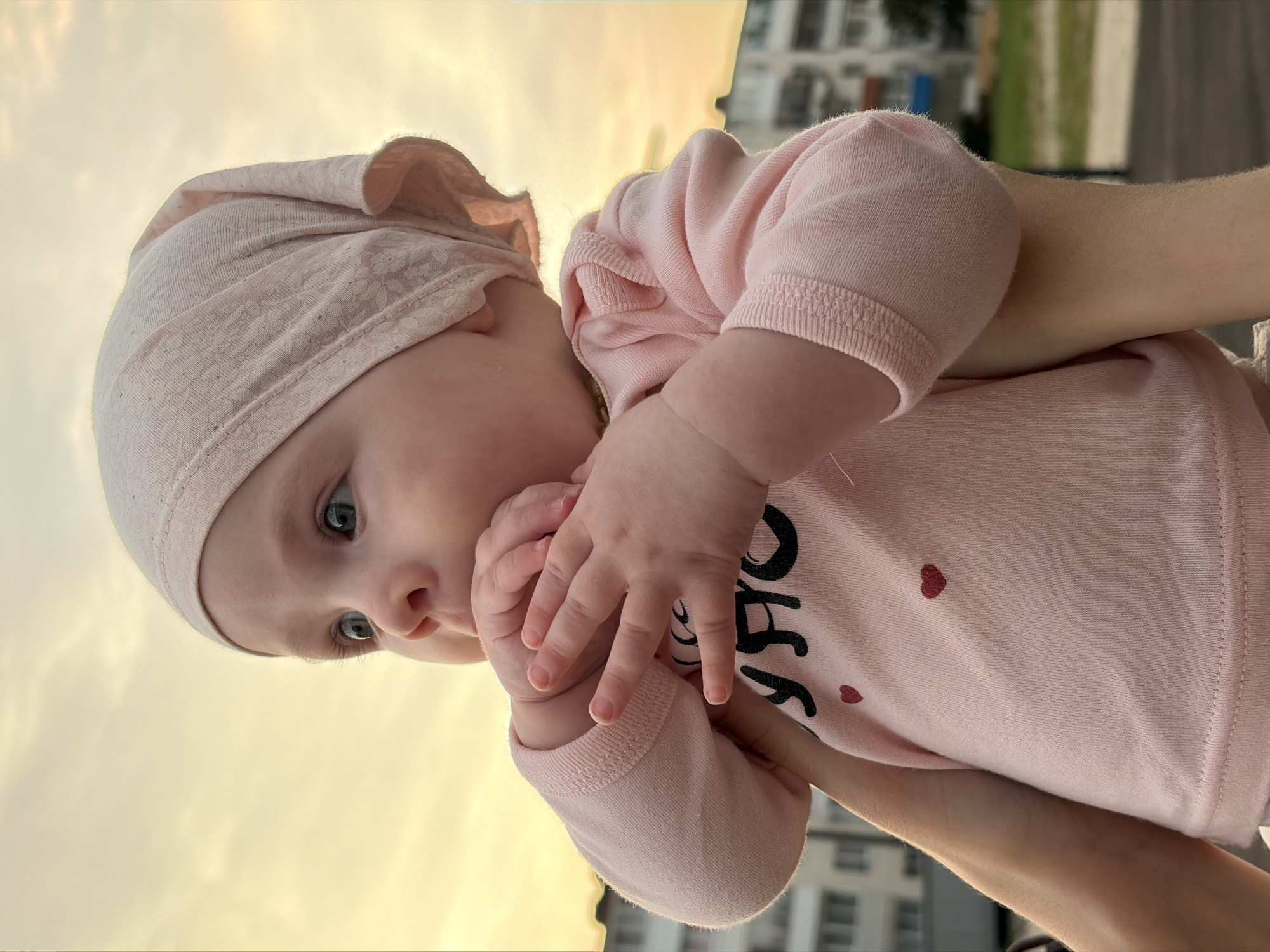 The image shows a close-up of a baby girl wearing a pink headscarf and a pink long-sleeved shirt with two small red hearts on it. Her hands are raised to her mouth. The background suggests an outdoor urban setting with buildings and a hazy sky illuminated by the setting sun. The image appears to have been taken at ground level, with the camera angled slightly upwards. The location is identified as Noviy, Russia, based on the provided information.