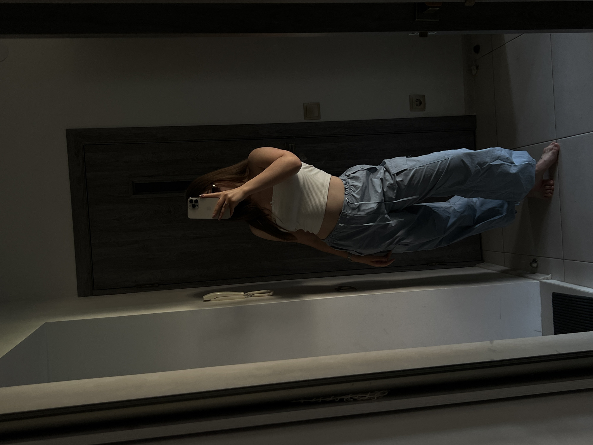 A full-body mirror reflection shows a young woman standing in a dimly lit interior, likely a hallway or entryway. She is taking a selfie with her smartphone, which is held horizontally. The woman is wearing a white crop top and loose-fitting light blue pants. Her hair is long and brown, and she is facing away from the camera, looking at her phone's screen.
The background features a dark wooden door with a horizontal accent panel. To the right of the door, a light-colored tiled wall is visible, with a power outlet on it. The floor appears to be tiled as well. In the foreground, a wide, reflective surface, presumably a large mirror, is present, capturing the scene. A light-colored telephone receiver rests on a surface below the door. The overall lighting suggests it is evening or the room is not well-lit. The image was taken in Ternopil, Ukraine.