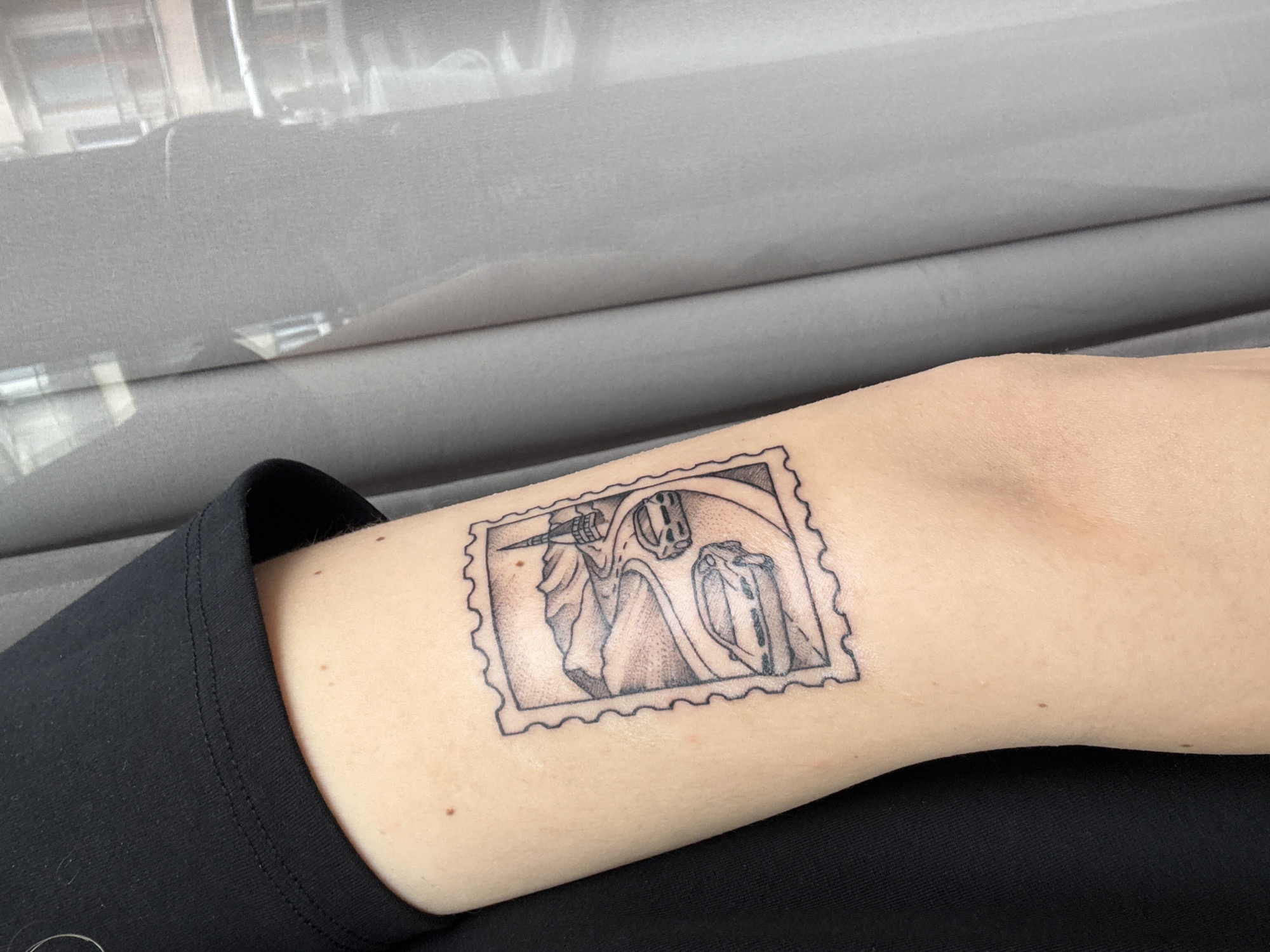 The image shows a tattoo on a person's inner arm. The tattoo is a black ink design resembling a postage stamp, featuring a grim reaper figure with a scythe next to what appears to be a piece of luggage. The person is wearing a black top. The background includes what appears to be a gray upholstered surface. The location in Varna, Bulgaria is not able to be determined from the image.