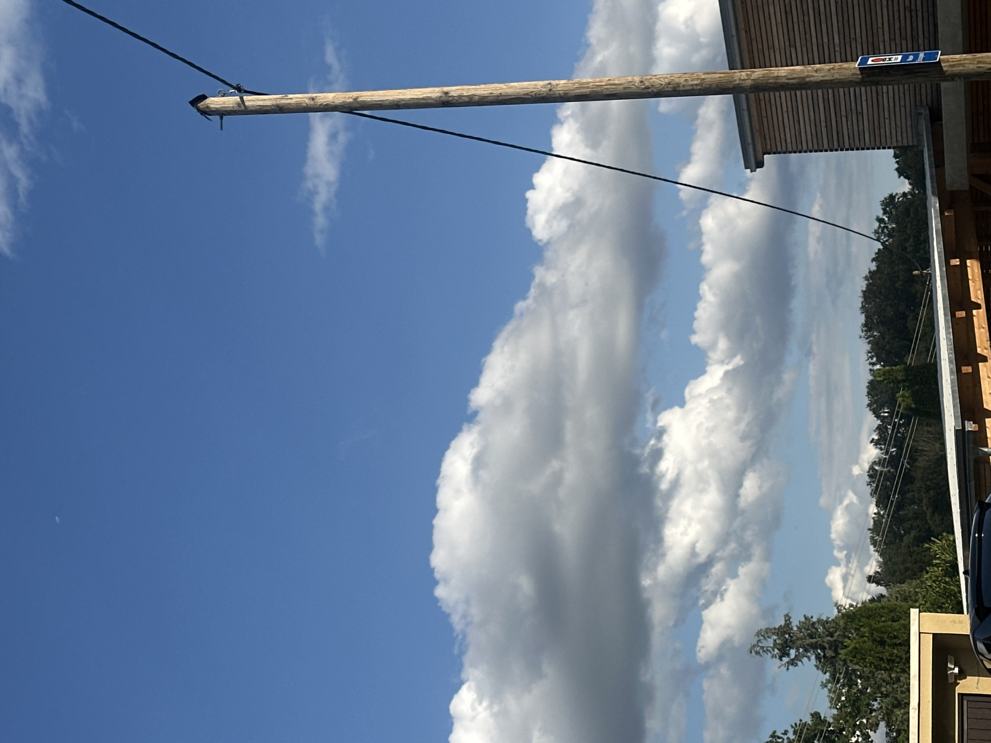 The image depicts a daytime scene with a clear blue sky featuring scattered white clouds. A thin crescent moon is visible in the upper left portion of the sky. A wooden utility pole stands prominently on the right side of the frame, with a black cable running diagonally upwards from the lower left to the top of the pole. At the base of the pole, a blue sign with white lettering and a red symbol is affixed. To the right of the pole, a modern building with horizontal wooden siding is partially visible. In the background, a dense line of trees stretches across the horizon. In the lower left, parts of a building with a beige facade and a dark roof are visible, along with the rear of a dark-colored car. The scene appears to be set in a suburban or semi-rural area. The presence of the building with wooden siding and the specific style of the signage suggests a location in Günzburg, Germany.