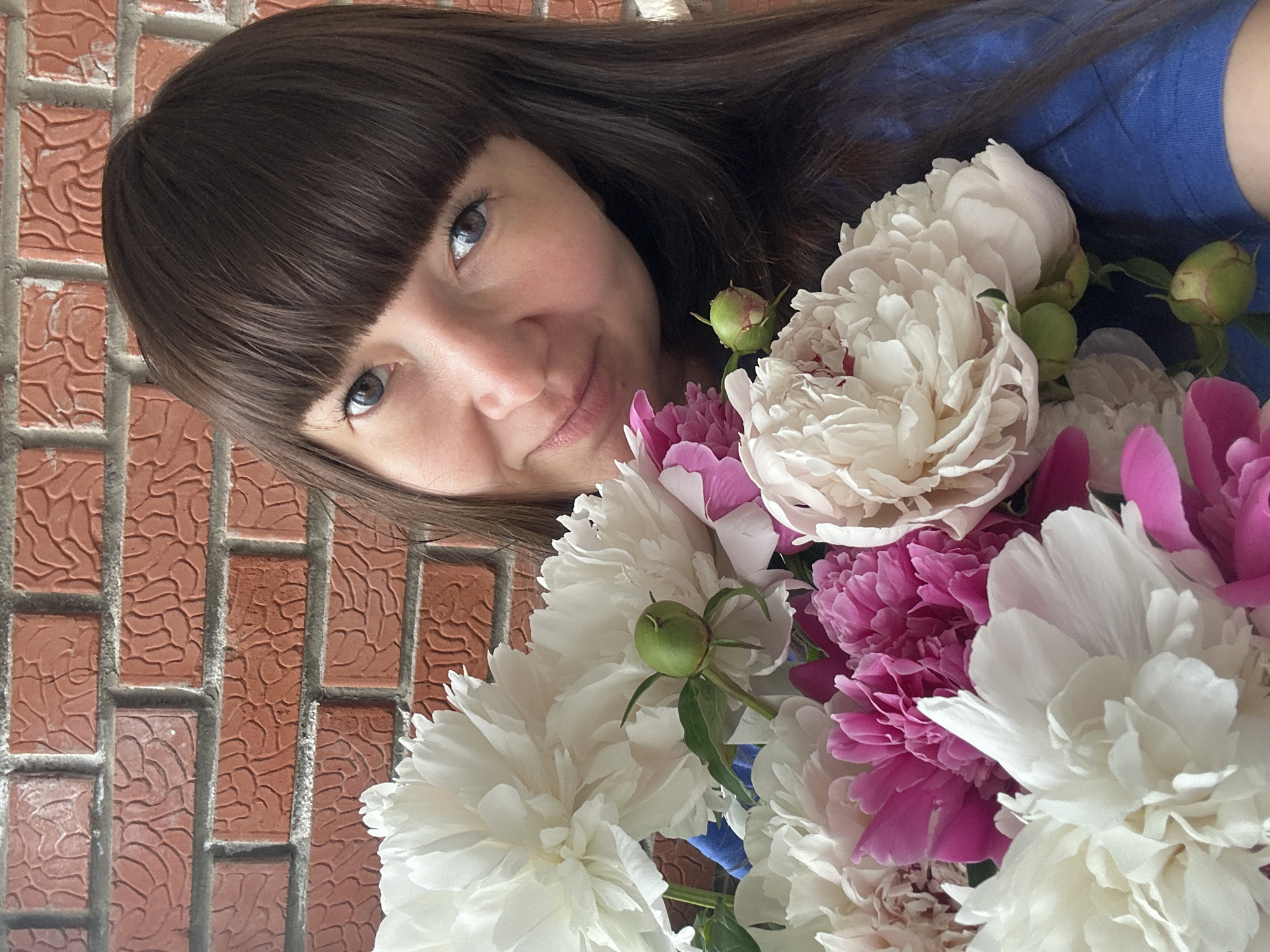 A woman with brown hair and bangs is posing with a bouquet of white and pink peonies. She is wearing a blue shirt and standing in front of a brick wall. The location is likely Chaikovsky, Russia, although there is no explicit confirmation within the image itself.