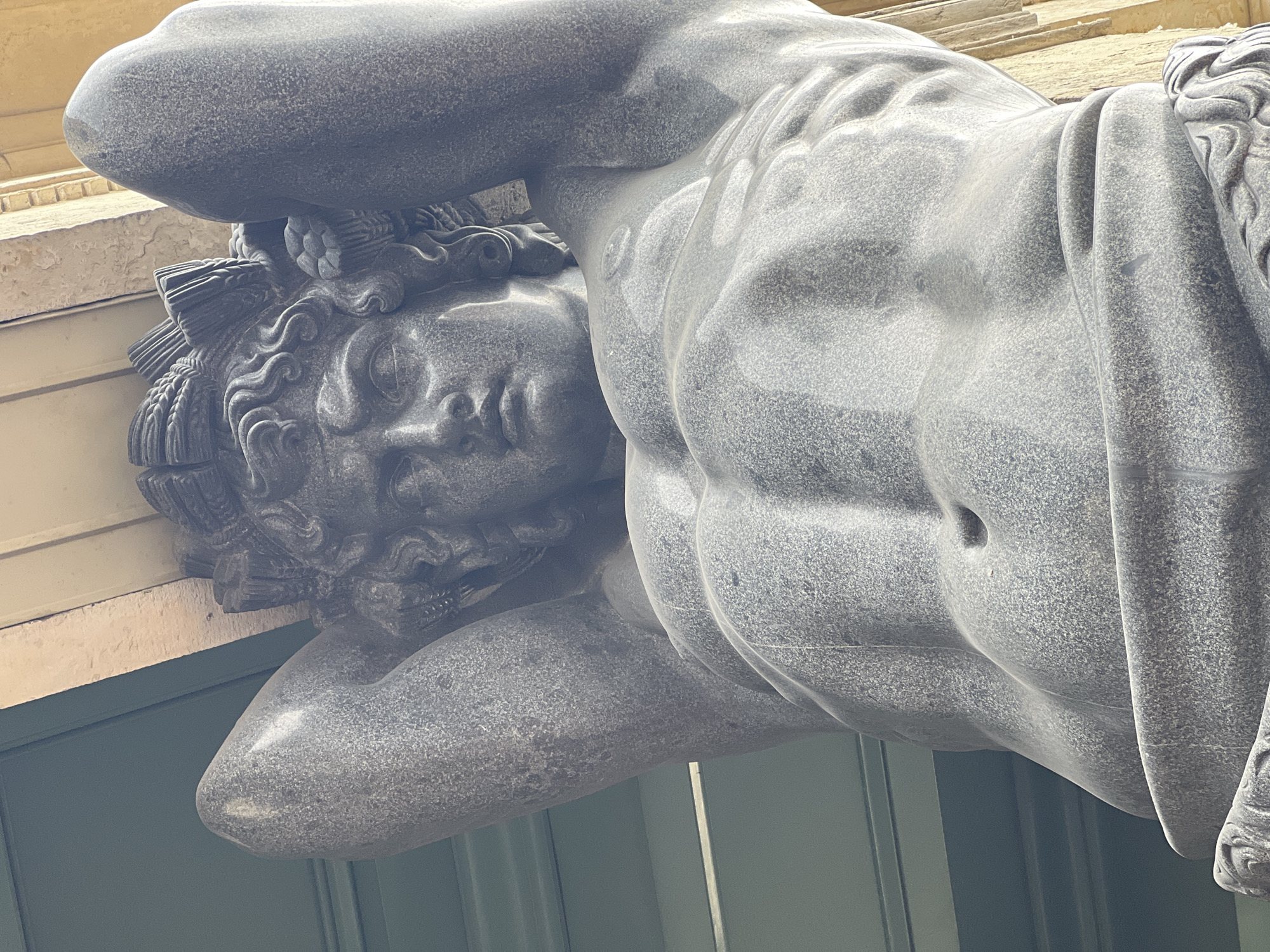 The image shows a close-up, upward-angled view of a large, grey granite statue of a muscular male figure. The statue is likely a caryatid or atlas, holding up an architectural element above its head with its hands. The figure has sculpted abdominal muscles and appears to be wearing a headdress with decorative elements. The background consists of a green, paneled surface and a beige, textured wall. This statue is located on the New Hermitage Museum on the Palace Embankment in Saint Petersburg, Russia.