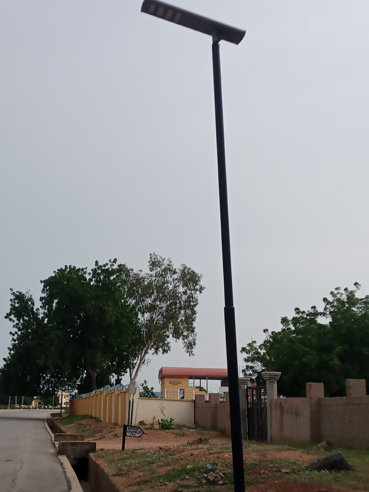 This is a medium shot, eye-level, outdoor, daylight photograph of a street scene in Jalingo, Nigeria. A tall, black, solar-powered street lamp with a rectangular light fixture dominates the right side of the frame, casting no discernable shadow. To the left of the street lamp, a paved road curves away from the viewer. Along the right side of the road is a concrete drainage channel. Beyond the drainage channel, a dirt path leads to a low, yellow wall topped with barbed wire. Behind the yellow wall, a building with a red roof is visible. A signpost on the path points to the right, indicating "ADONAI CHAPEL". Further right, a more substantial wall made of concrete blocks with decorative pillars is visible, with a wooden gate set within it. Trees with dark green foliage are present in the background. The sky is overcast with a pale gray hue. No people are visible in the image.