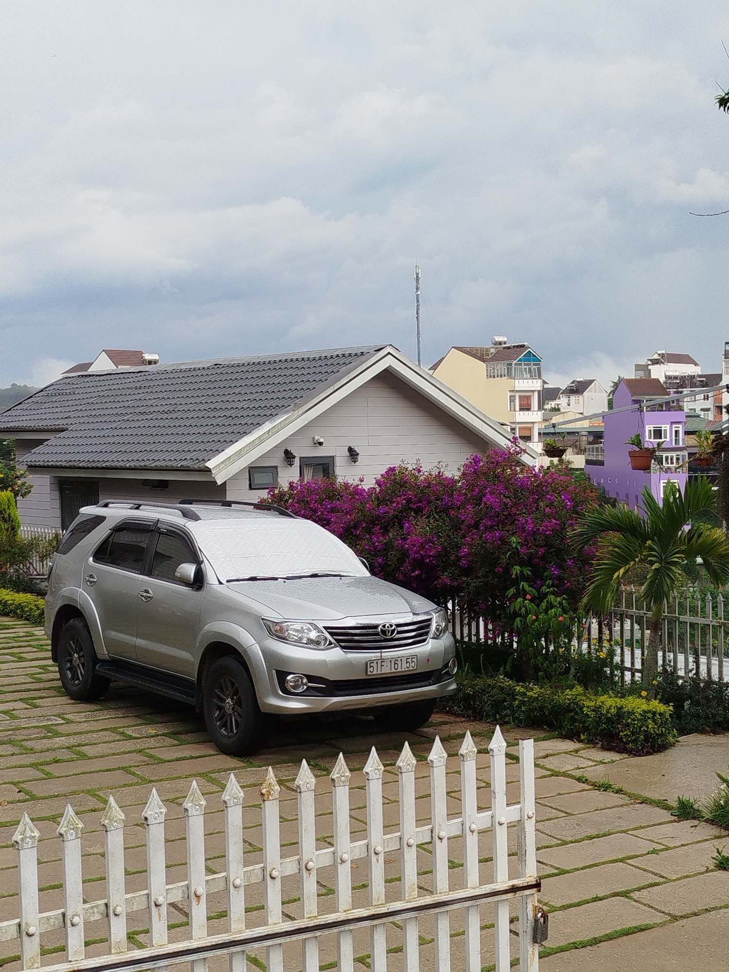 A silver Toyota Fortuner is parked in front of a light-colored house with a gray roof in Da Lat, Vietnam. A silver sunshade is on the windshield. Purple bougainvillea and a small palm tree are between the house and a white picket fence. Paving stones cover the driveway. In the background, buildings of varying colors and heights are visible, and a cell tower rises above them. The sky is overcast.