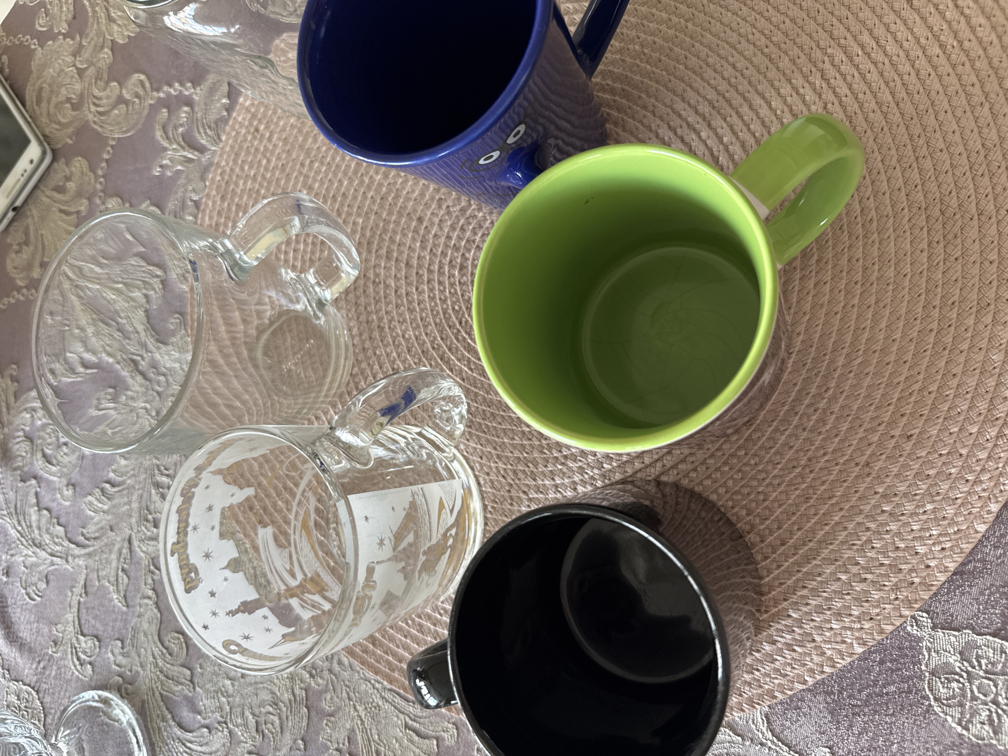 A high-angle, full shot shows four mugs on a table. The mugs are of varying colors and designs. A clear glass mug with a silhouette of a city and stars is on the left. Next to it is another clear glass mug with a handle. In the foreground, a black mug is on the left, and a bright green mug is on the right. A dark blue mug with a face design is positioned behind the green mug. The mugs are placed on a woven placemat, which rests on a patterned tablecloth. A phone can be seen in the background. The scene appears to be indoors, possibly in a home or cafe.