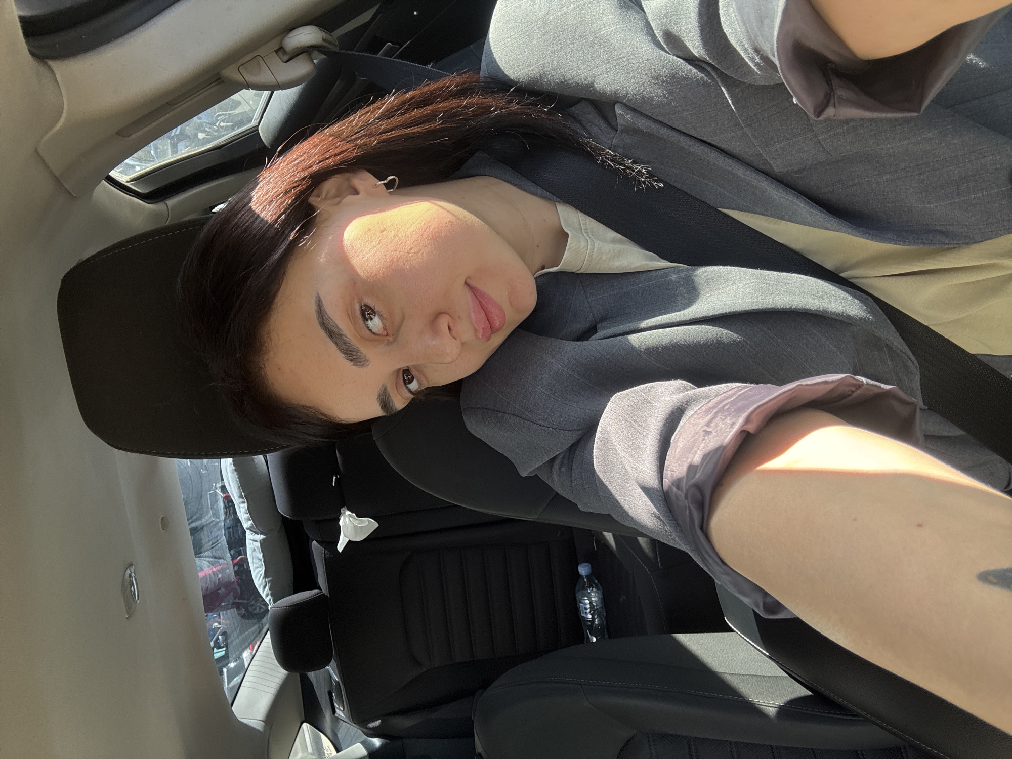 A woman in a car, wearing a gray blazer and a seatbelt, is taking a selfie. She has dark brown hair and is leaning her head to the side. The car has black leather seats, and a water bottle is visible on the passenger side floor. The car has a sunroof. The city of Tbilisi, Georgia cannot be determined from the image.