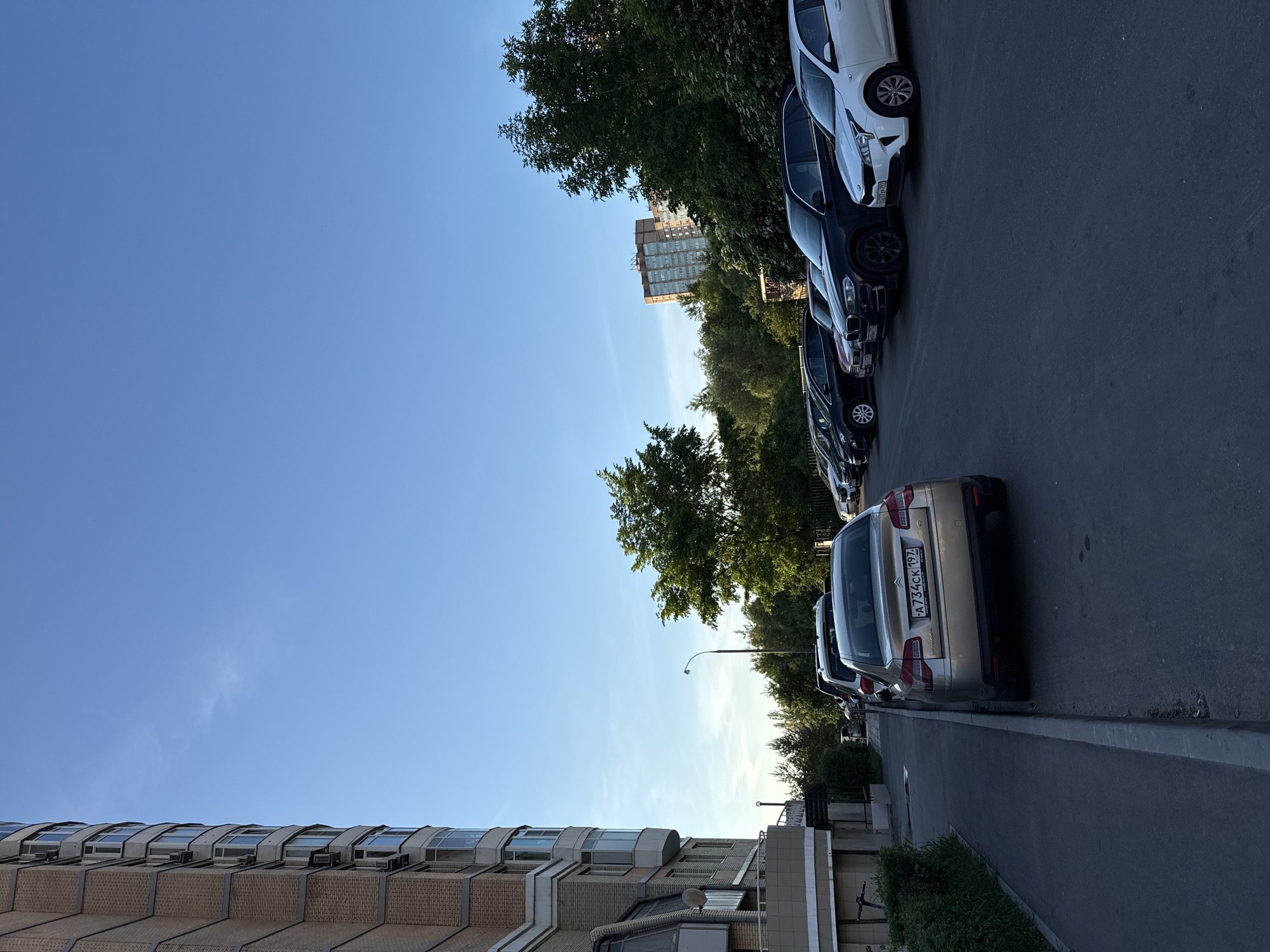 This outdoor image, taken during daytime, depicts a street scene with parked cars, buildings, and abundant greenery under a clear blue sky.
A row of cars is parked bumper-to-bumper along the right side of a paved road. The closest vehicle is a light gold/beige sedan, which has a Russian license plate clearly visible: "A734CK 197 RUS". Behind it, several dark-colored sedans are parked, followed by a white SUV or hatchback at the far end of the line. A partial license plate "O308TH 777" is visible on one of the dark cars.
To the left of the road, a curb delineates a narrow strip of grass, beyond which stands a multi-story building. This building has a light-colored brick facade and a distinctive architectural style with curved window sections along its upper levels, possibly featuring skylights or air handling units. Further back and among the trees, a street lamp is visible.
Lush green trees line the street, forming a dense canopy behind the parked cars and extending into the background. In the distance, beyond the trees, another multi-story building with numerous windows can be observed. The sky overhead is a vibrant, cloudless blue, suggesting fair weather and good visibility. The lighting indicates it is daytime, likely mid-morning or late afternoon, as there are no harsh overhead shadows. No people or specific activities are visible, presenting a tranquil streetscape. A purple scooter is partially visible on the sidewalk in the bottom left of the image.