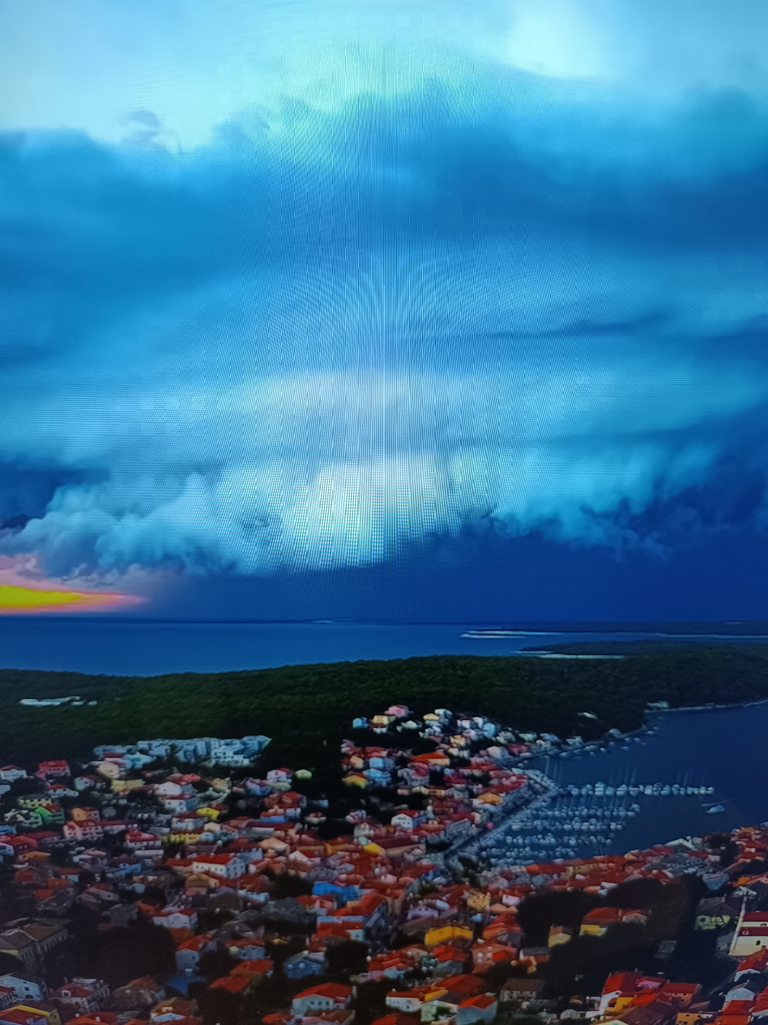The image depicts a coastal town with densely packed, colorful buildings situated on a hillside overlooking a body of water. The architecture suggests a Mediterranean or European influence, with red-tiled roofs and a variety of pastel and earth-toned facades. A marina with numerous sailboats is visible along the coastline. Above the town, a dramatic sky is filled with dark, ominous storm clouds, while a sliver of sunset or sunrise is visible on the horizon, casting a warm glow of yellow and pink. The foreground shows more of the town's rooftops and greenery. The image does not contain any specific elements that would place it in Taraz, Kazakhstan; instead, the visual cues suggest a different geographical region.