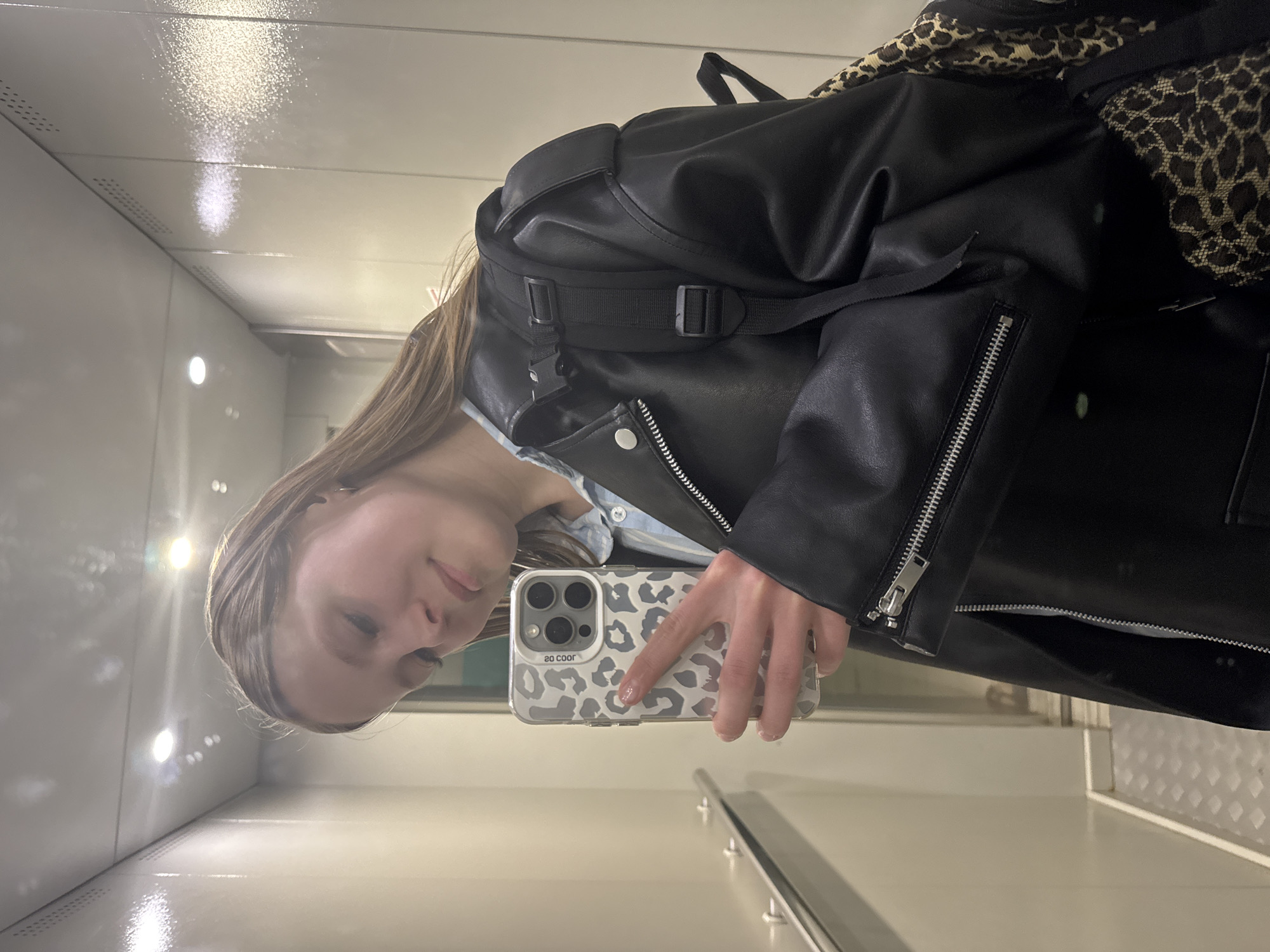A young woman takes a selfie in what appears to be an elevator or small enclosed space with shiny, white walls and ceiling. She is holding a cell phone with a leopard print case. She is wearing a black leather jacket over a light blue shirt, and has a backpack with a leopard print design slung over her shoulder. The location cannot be determined from the image, nor can a link to Chaikovsky, Russia.
