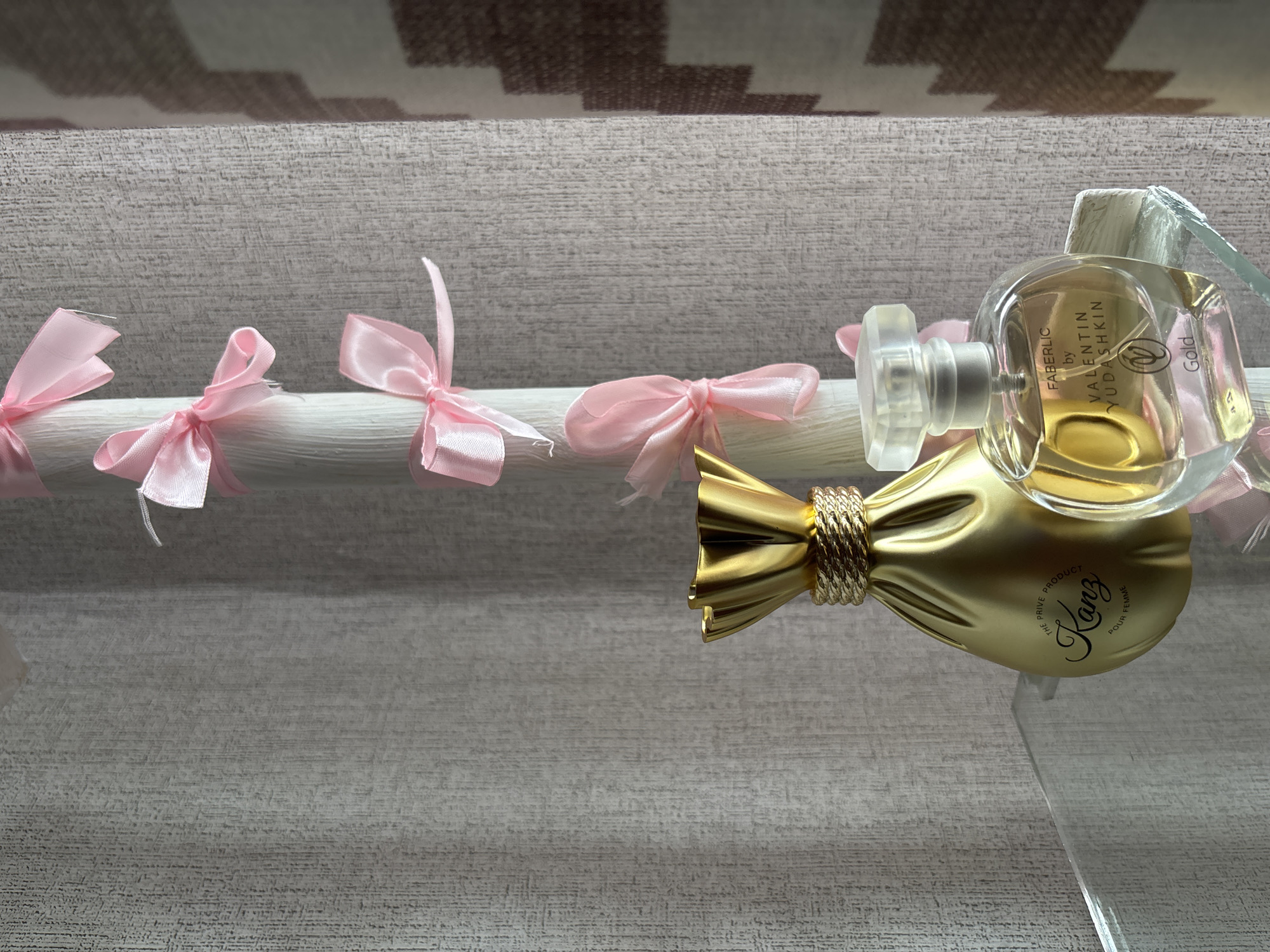This is a close-up, eye-level, indoor shot of two perfume bottles placed on a clear acrylic shelf against a textured grey backdrop. To the left is a gold-colored perfume bottle shaped like a pouch or coin purse with a gold rope-like detail around its neck. Text on the bottle reads "Kanz Pour Femme". To its right is a clear, round perfume bottle with a clear, faceted cap. Text on this bottle reads "FABERLIC by VALENTIN YUDASHKIN Gold". A white vertical object, possibly a pillar or display stand, is visible behind the bottles, adorned with several pink satin ribbons tied into bows. The overall lighting is soft and diffused. The image does not provide any specific information about the city of Inskoy, Russia, or its features; this detail is external information.